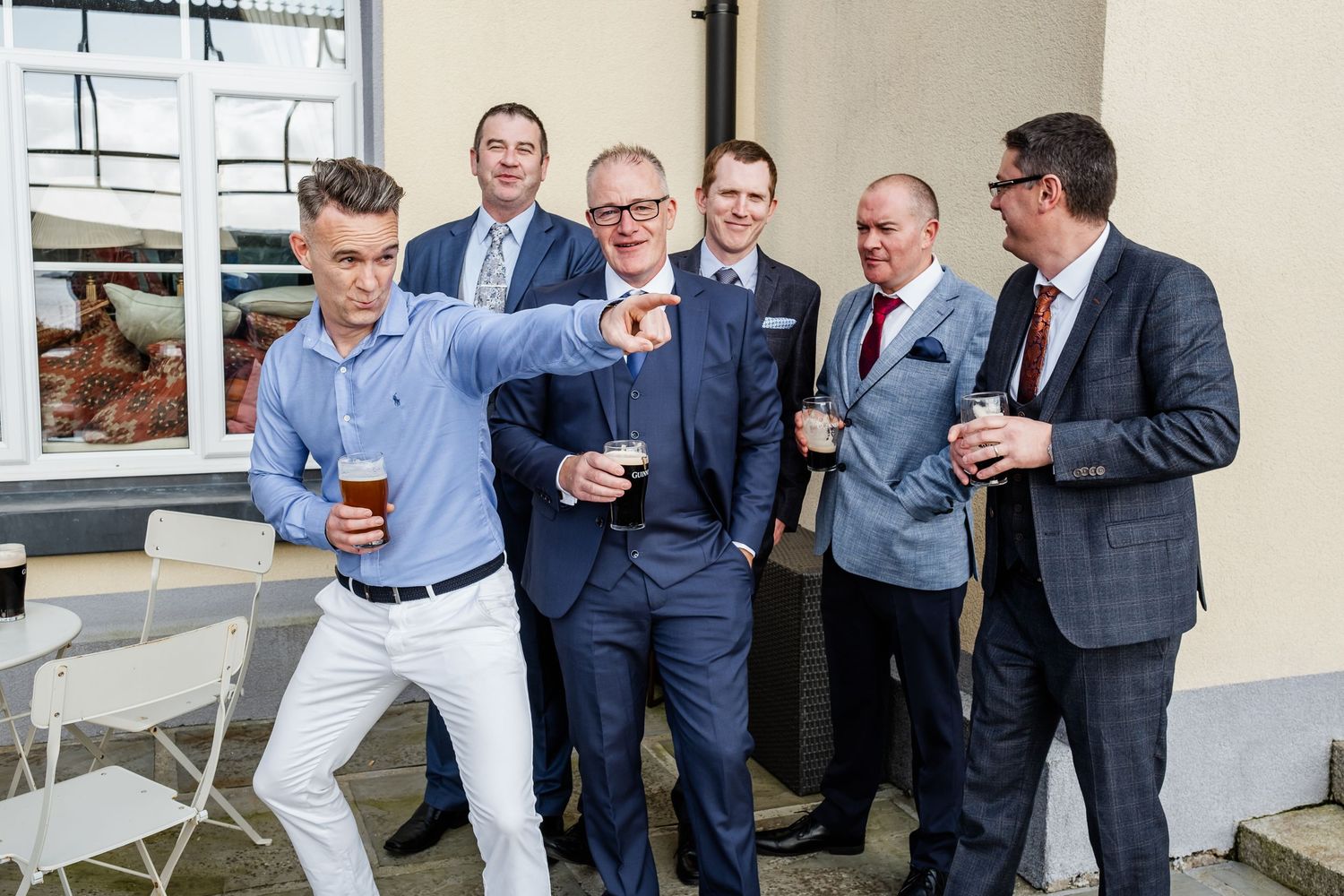 Group of well-dressed men in suits socializing outdoors, holding drinks at a formal wedding celebration event.