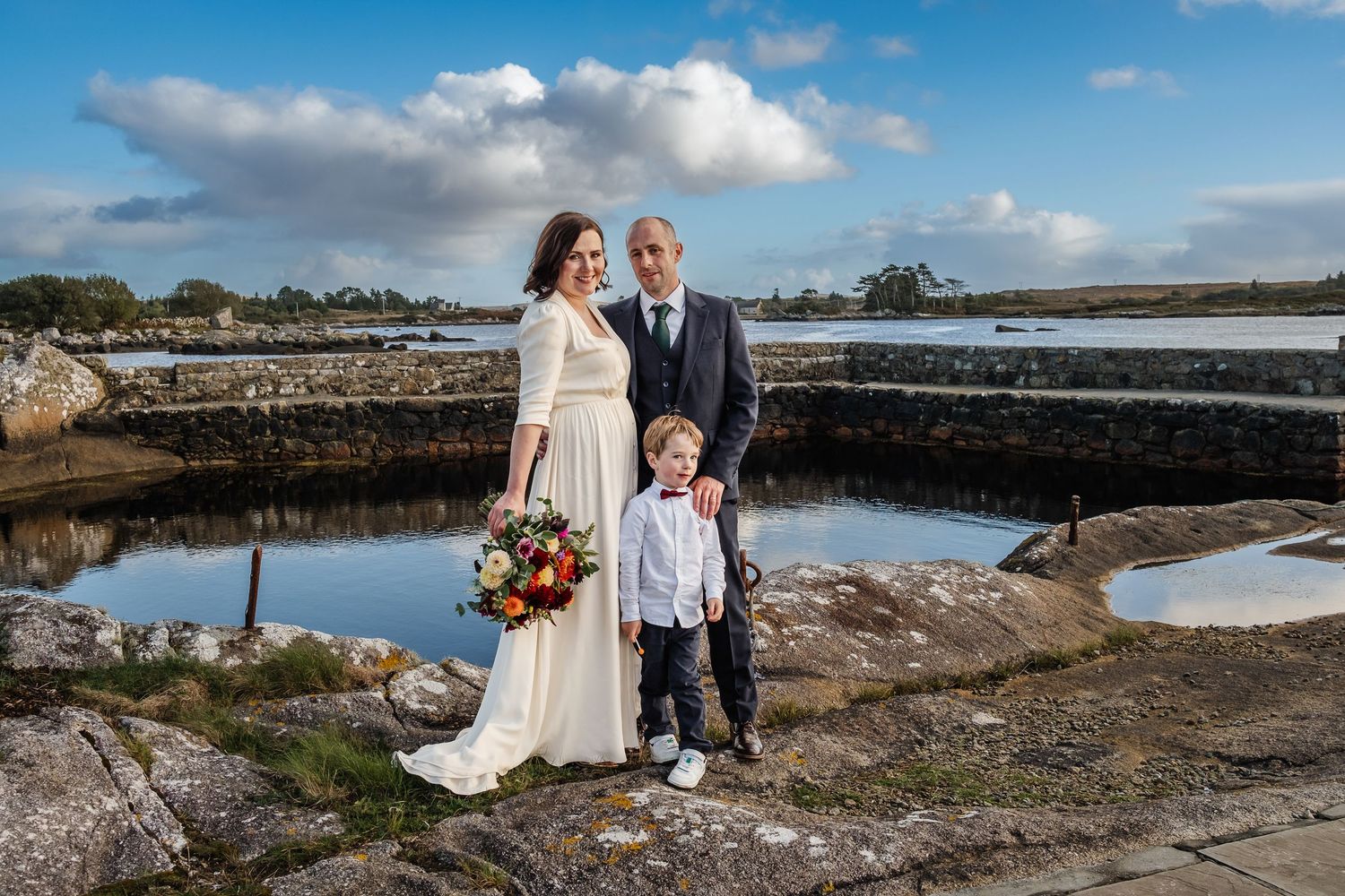 Wedding couple with young boy poses by a scenic coastal harbor on a sunny day, bride holding a colorful bouquet.