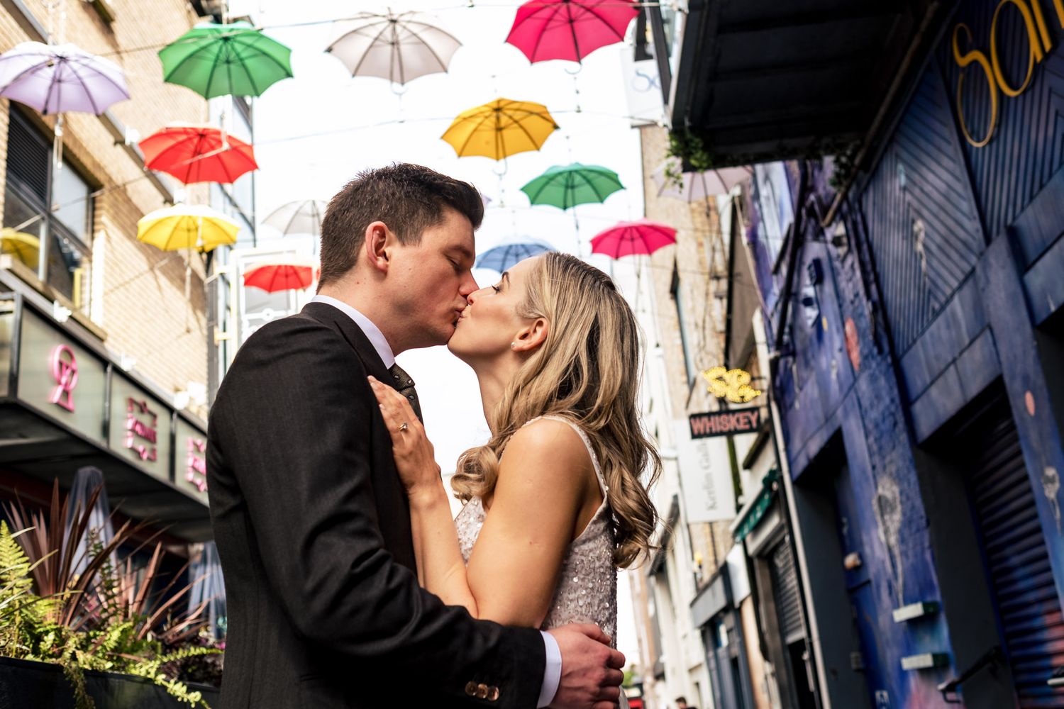 Colourful umbrellas hanging overhead create a festive canopy on an urban street with kissing couple.
