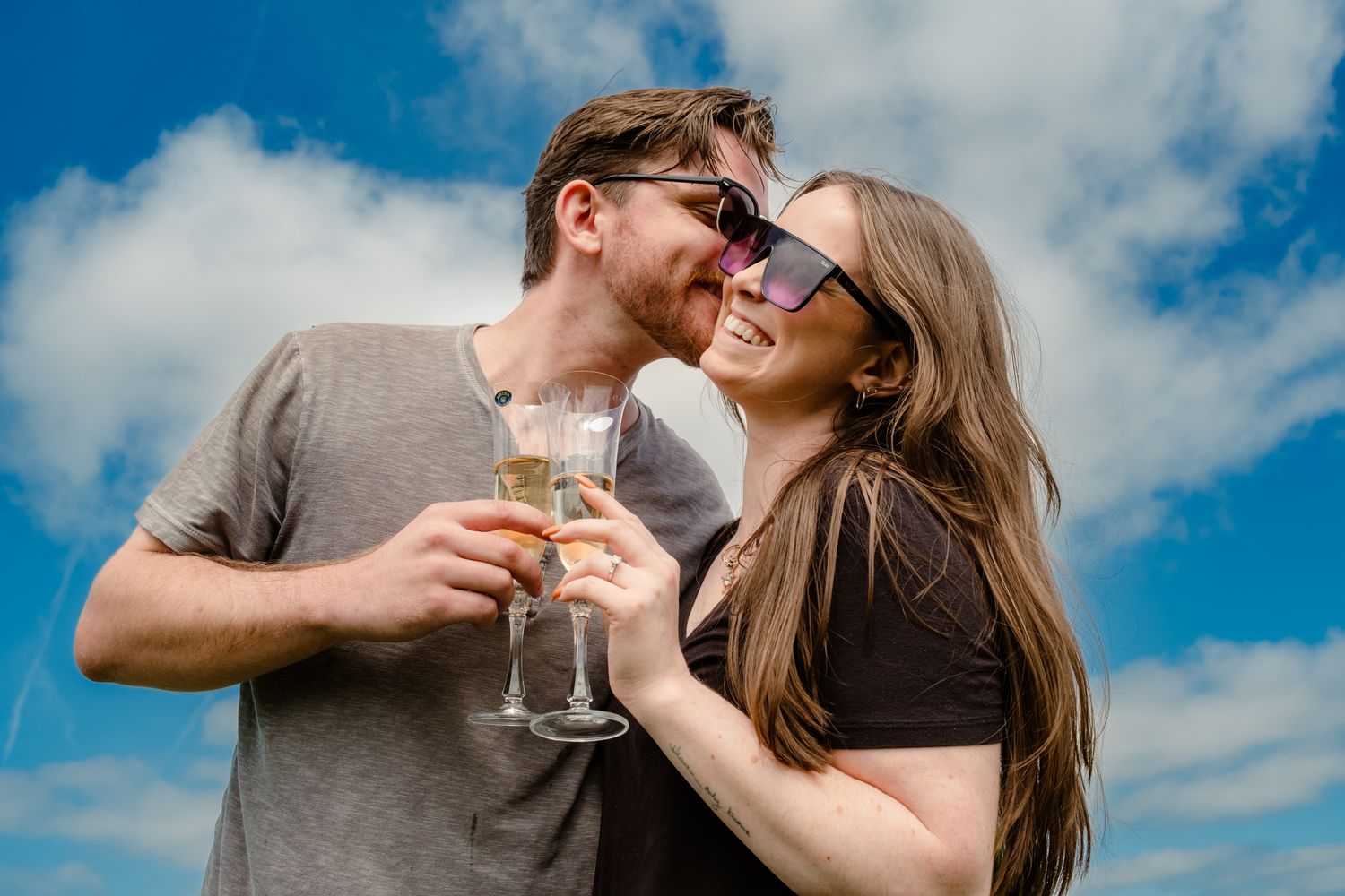 A joyful couple toasting with wine glasses against a bright blue sky with clouds.