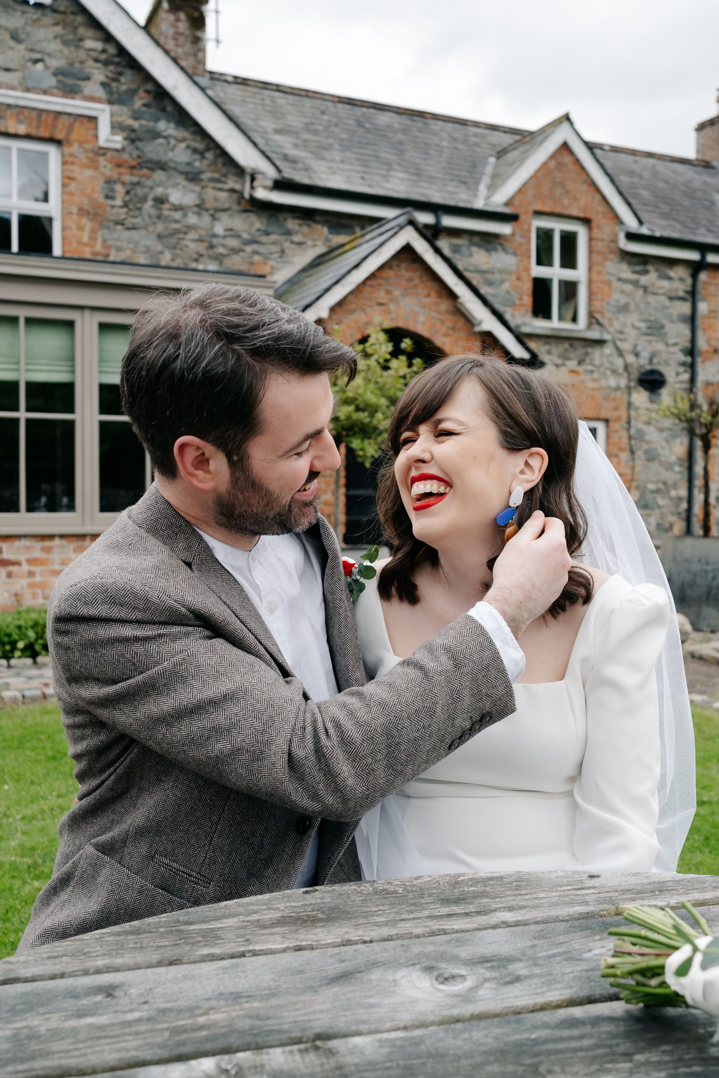 A smiling groom in a grey tweed suit adjusts his bride's earring outdoors near a stone building on their wedding day.