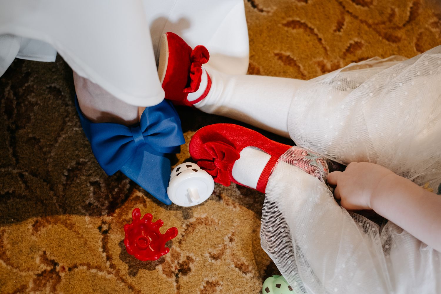 Close-up of blue bow wedding shoes and red doll shoes on a floral carpet, bride getting ready.
