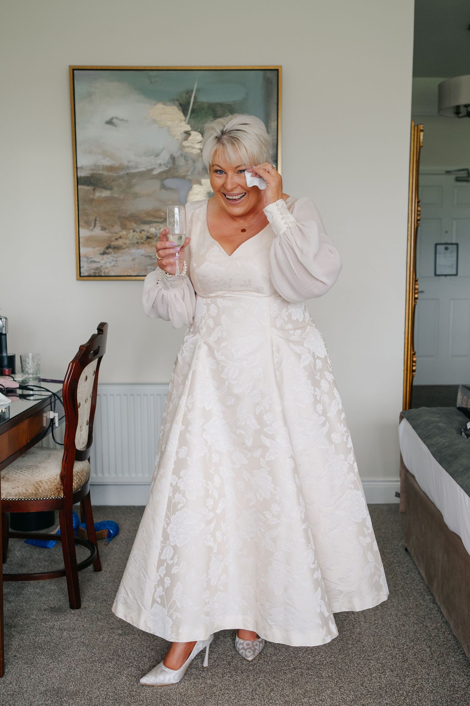 Emotional bride in white lace tea-length wedding dress holding champagne glass, wiping tears of joy in hotel room.