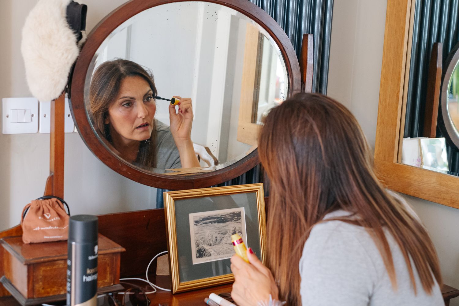 Woman applying mascara at a vintage wooden vanity mirror on a dressing table with framed photos.