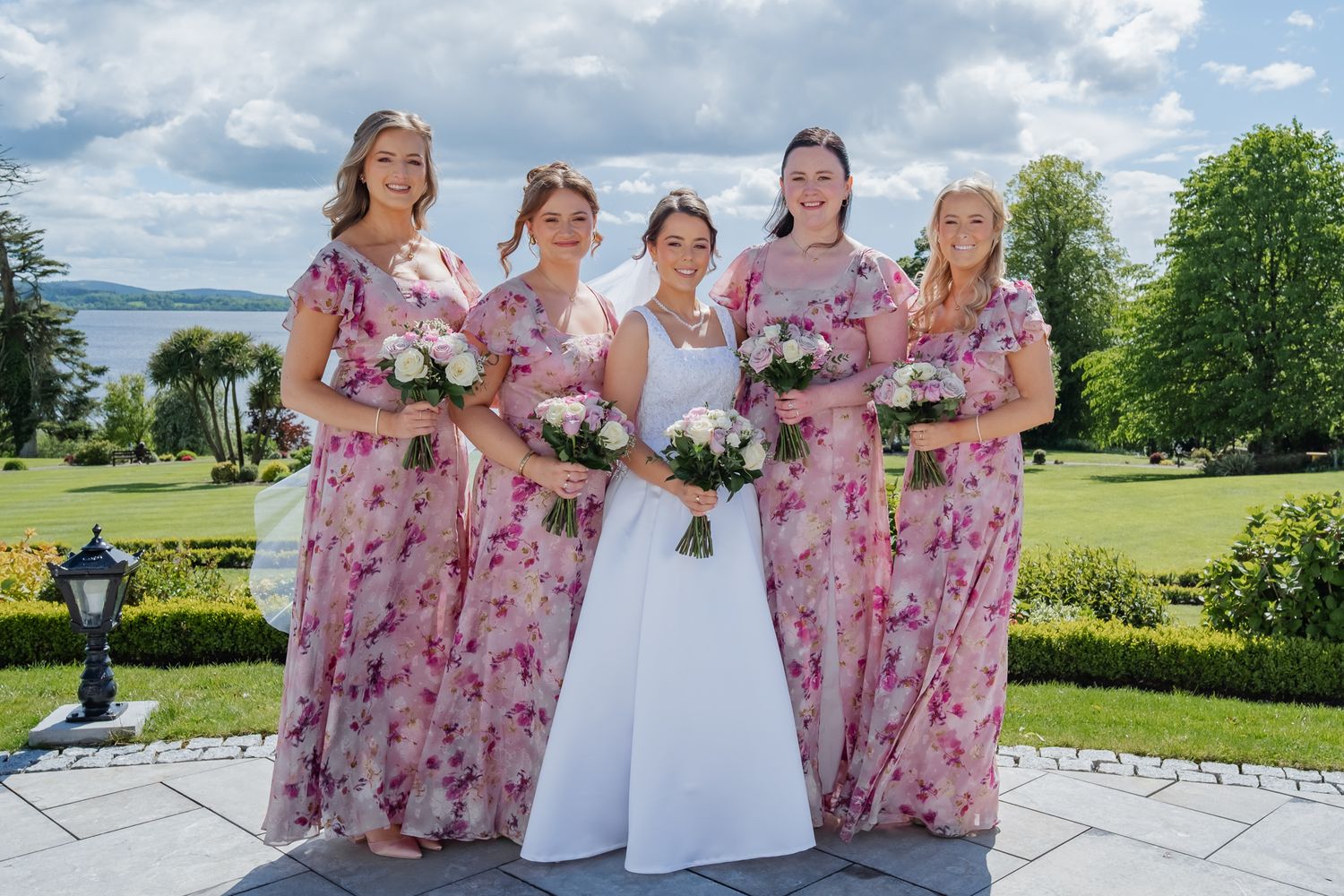Bride in white gown poses with four bridesmaids in pink floral dresses holding bouquets in a lush garden setting.