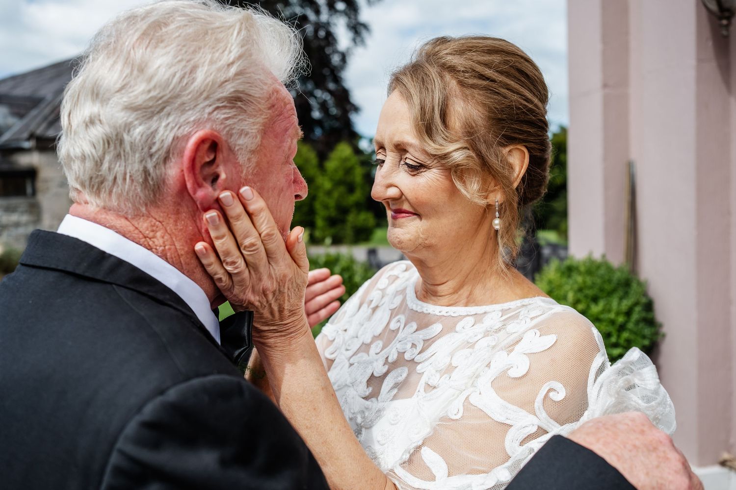 Elderly bride in white lace gown tenderly cups groom's face during an emotional outdoor wedding ceremony.