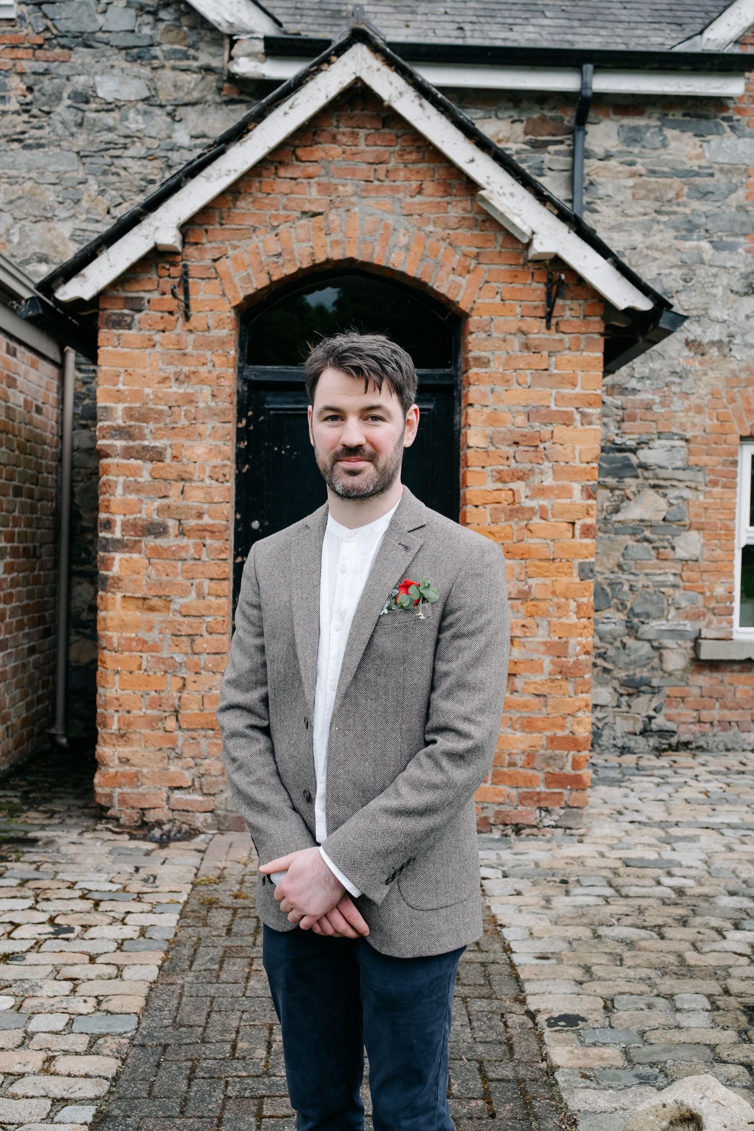 A groom in a grey tweed blazer with a floral boutonniere stands smiling before a rustic brick building.
