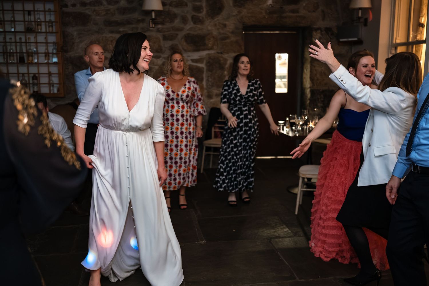 Guests dancing joyfully at a wedding reception in a rustic stone venue, woman in white dress leads the celebration.