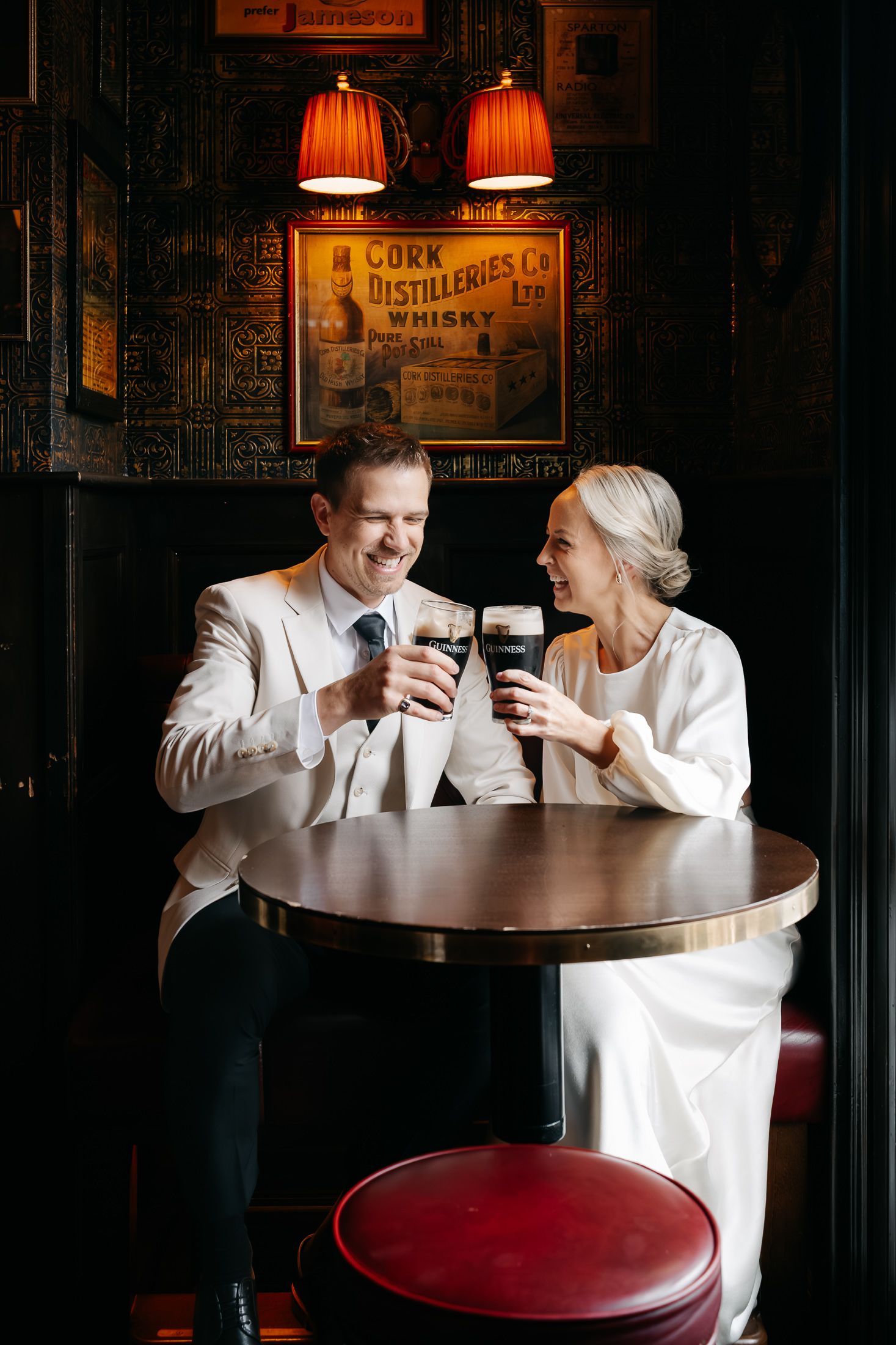 Newlyweds toast with Guinness pints at a dark Irish pub, dressed in wedding attire, smiling joyfully.