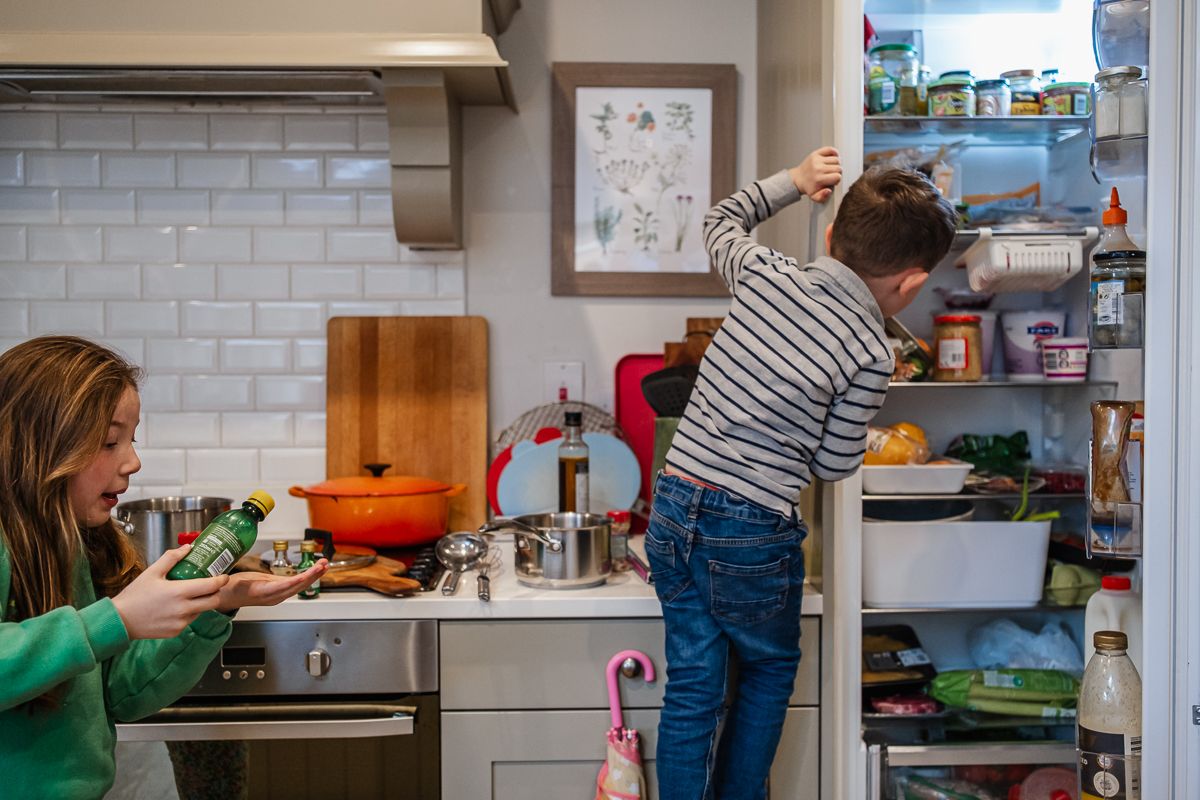 Two children in the kitchen, one looking in refrigerator while other helps with meal preparation.
