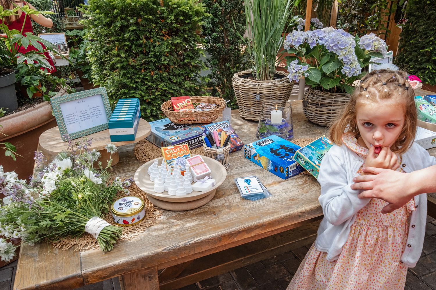 Young girl in pink dress stands beside a rustic wooden table with flowers, crafts, and activity supplies outdoors.