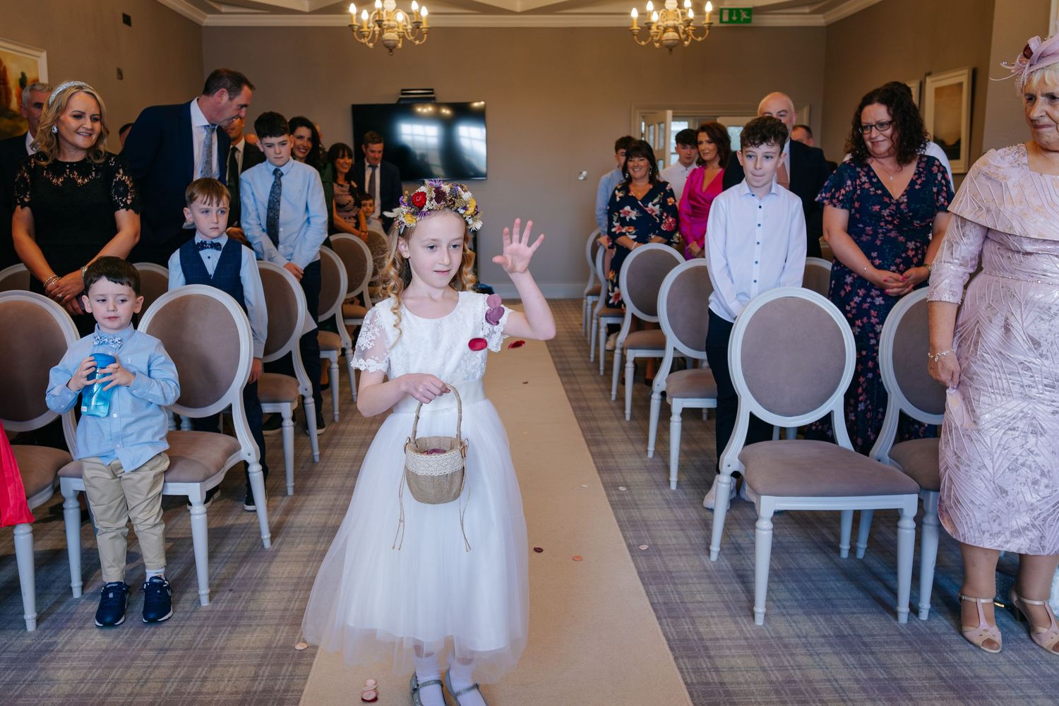 Flower girl waving while walking down the aisle at a wedding ceremony, with guests seated on either side.