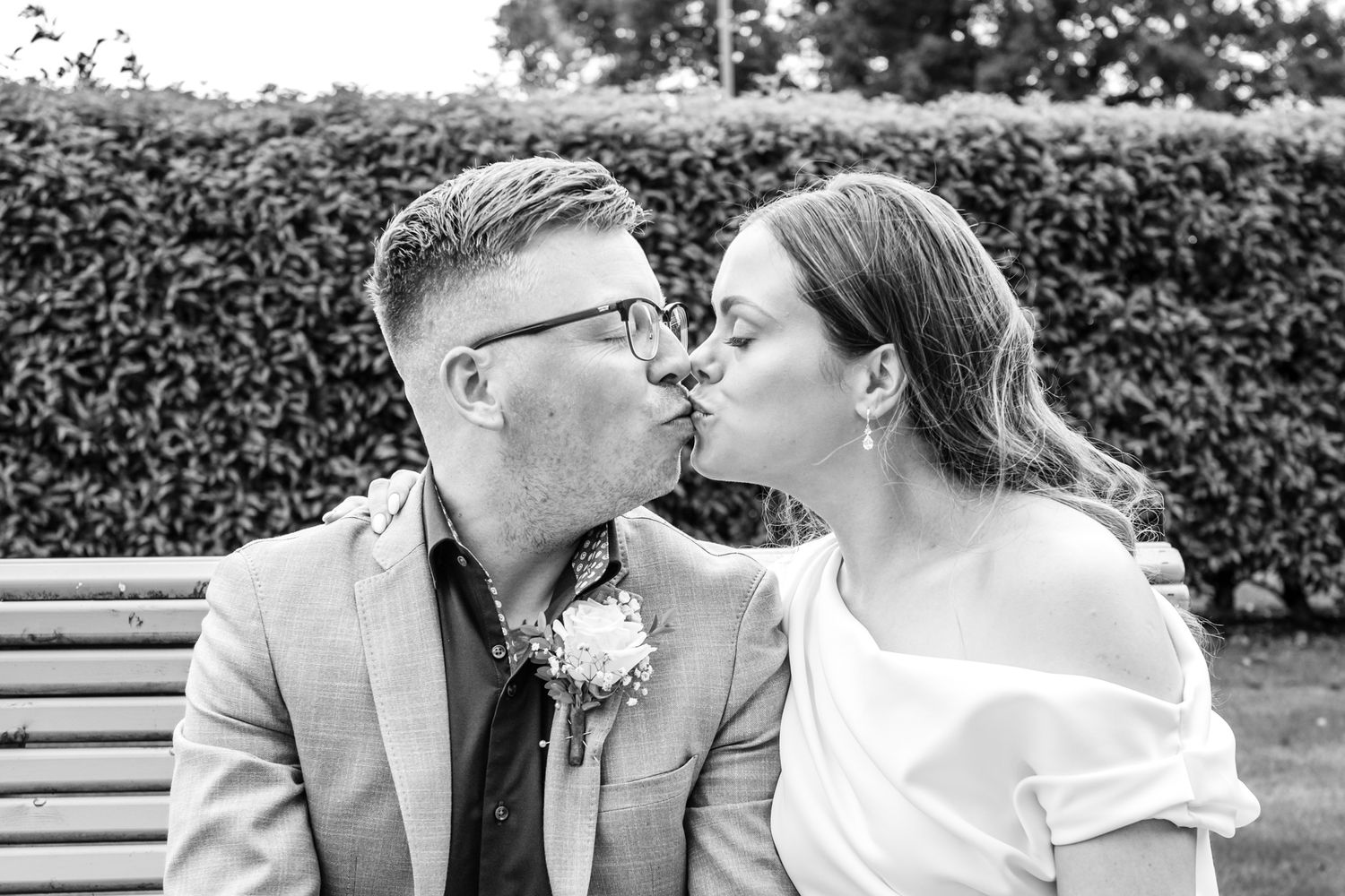 Black and white romantic moment captured on a park bench with hedge backdrop.