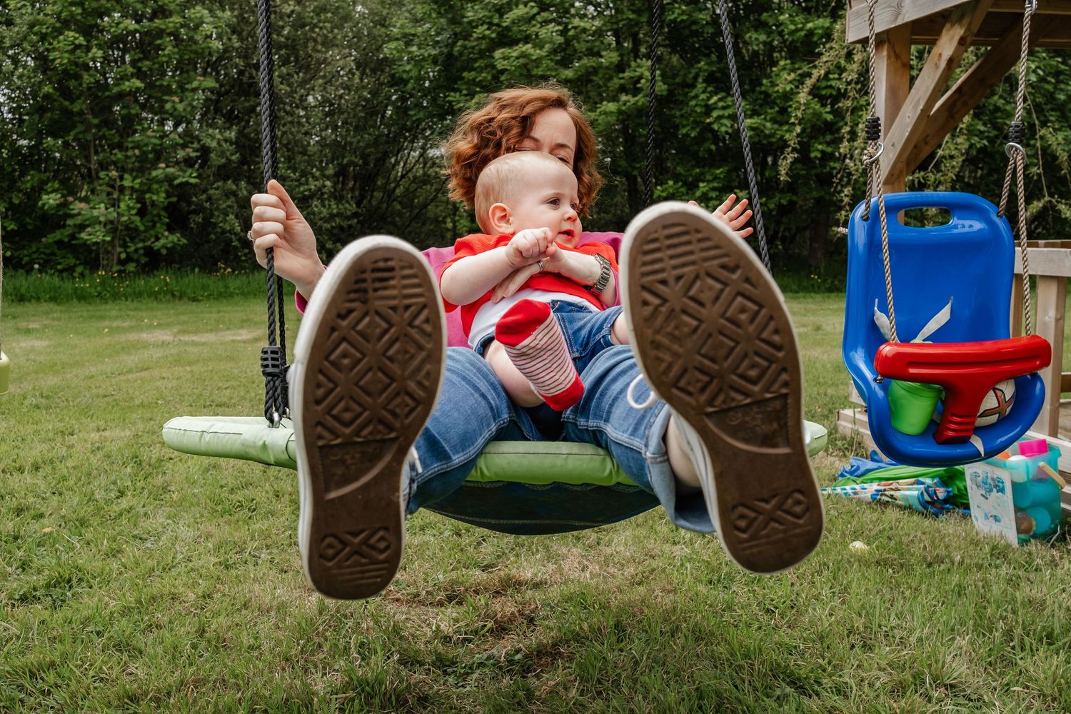 Child in red clothing swinging outdoors with feet prominently displayed against garden background.