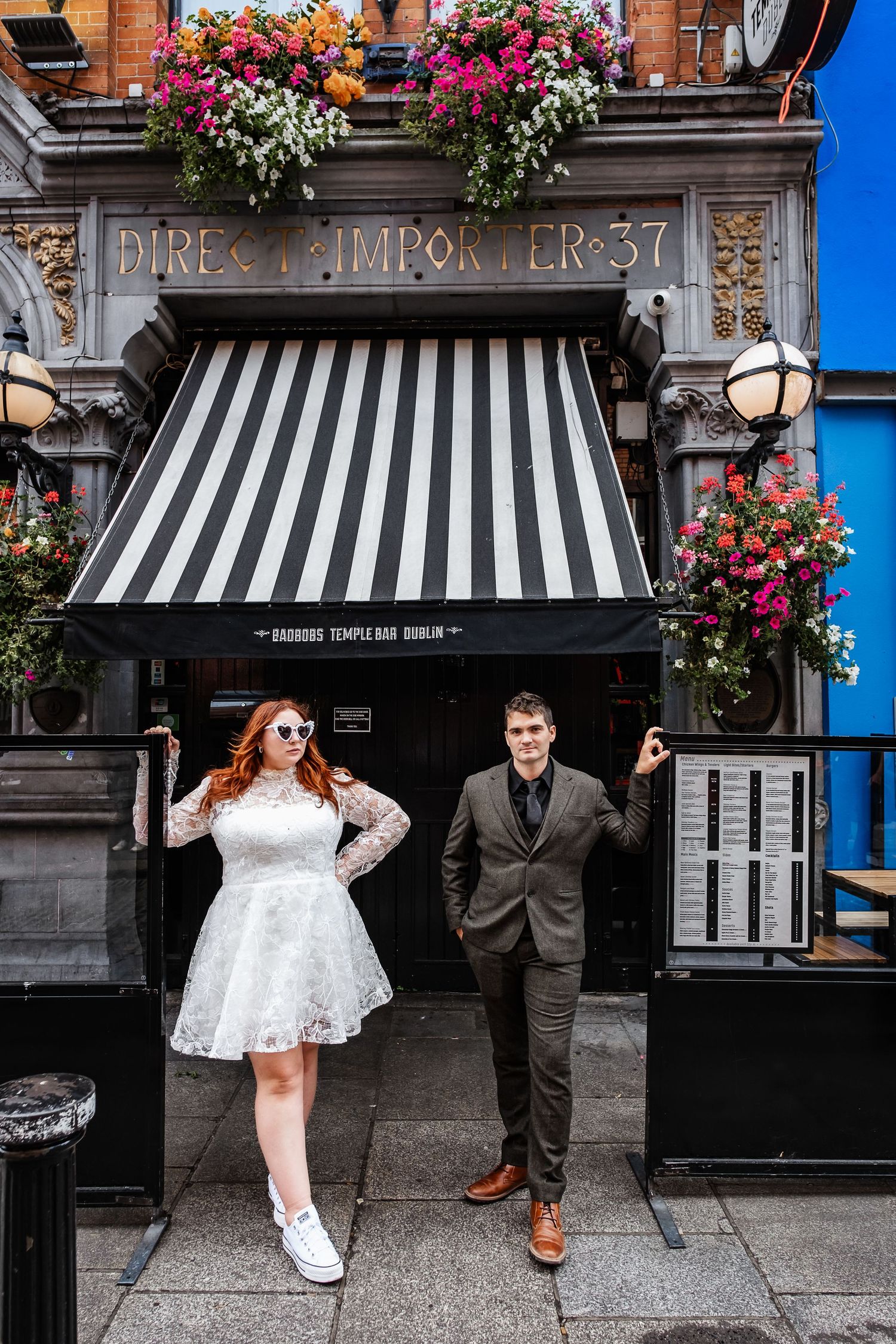 A couple poses playfully outside a pub with hanging flower baskets and striped awning.