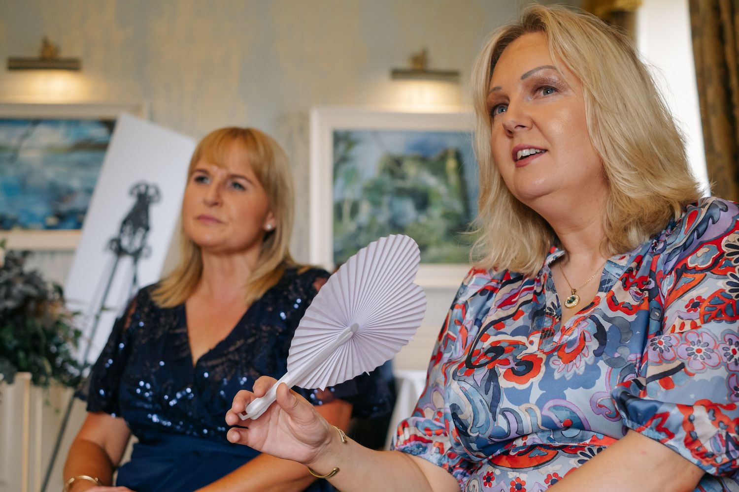 Two women seated at wedding reception, one holding white paper fan.