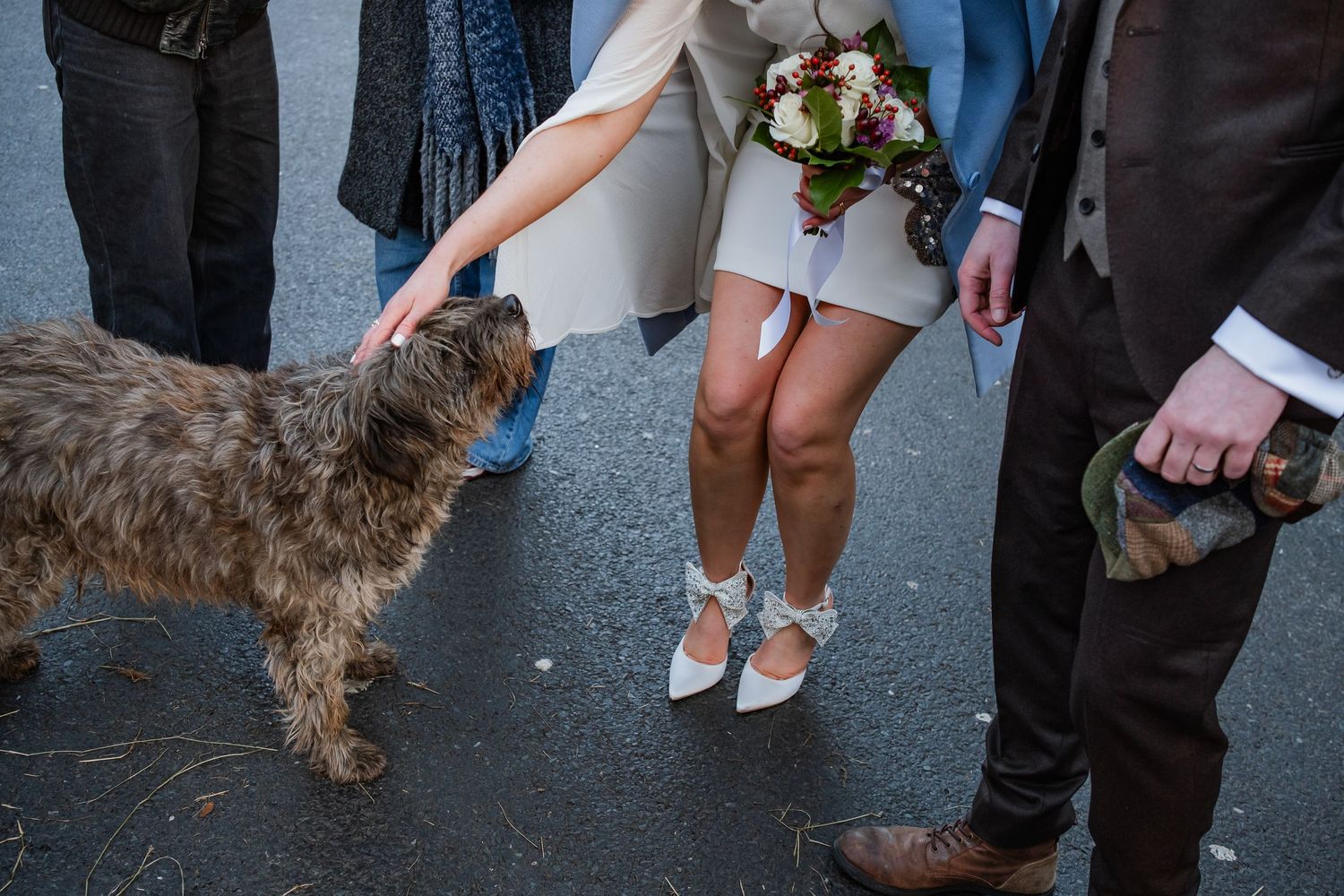 Bride in white mini dress and heels holds bouquet while petting a dog at an outdoor wedding celebration.