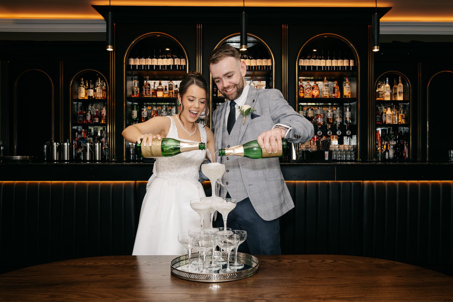 Bride and groom pouring champagne tower at elegant dark bar with bottle-lined shelves at wedding reception.