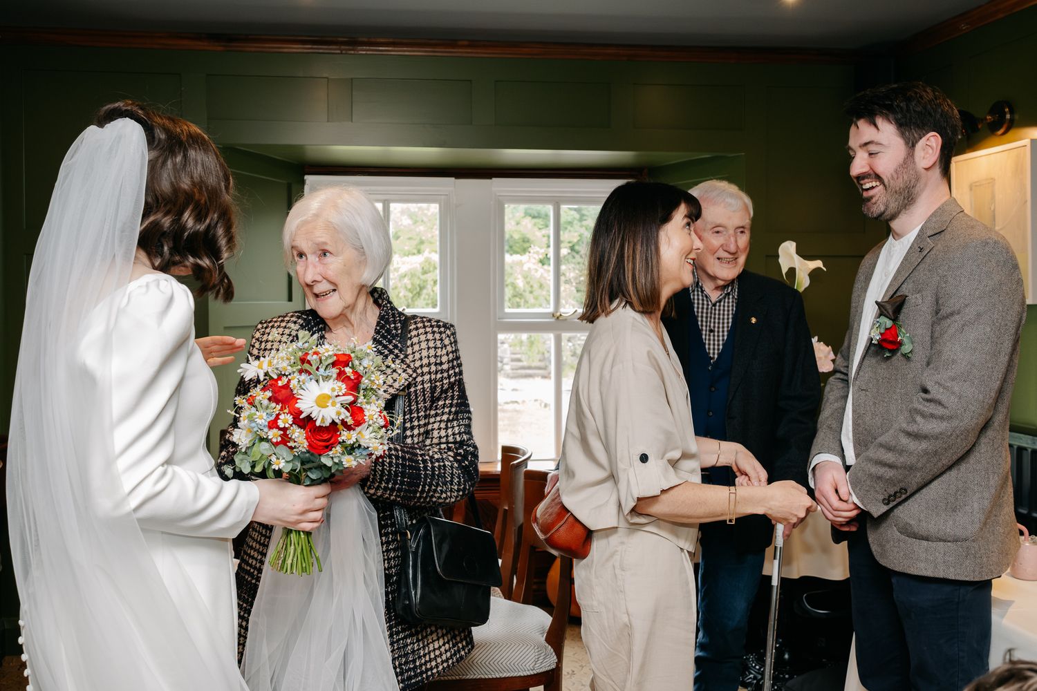 Elderly woman receives bridal bouquet of red and white flowers at an intimate wedding reception gathering.