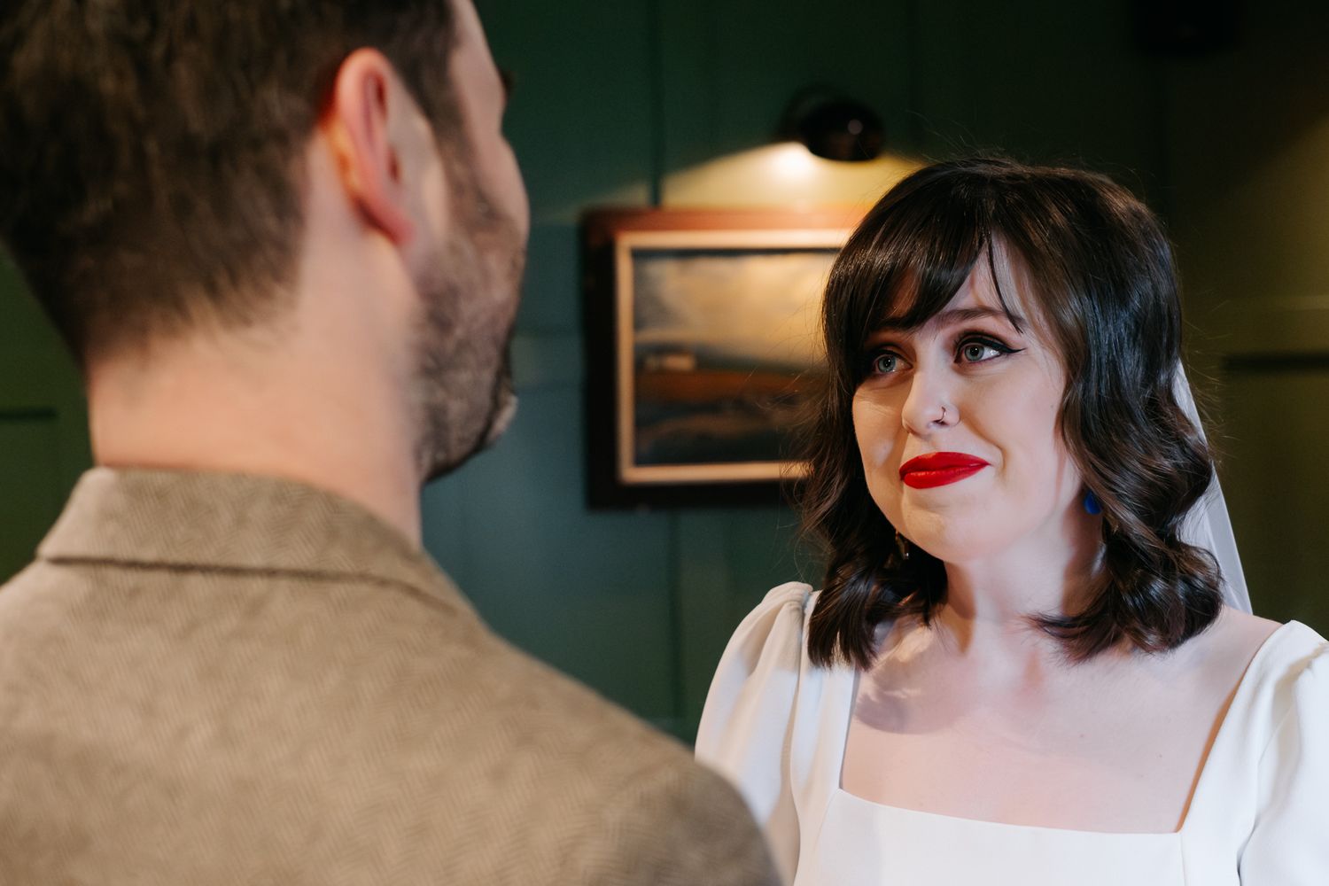 A woman in a white dress with red lips gazes at a man in a gray jacket inside a dark-walled room.