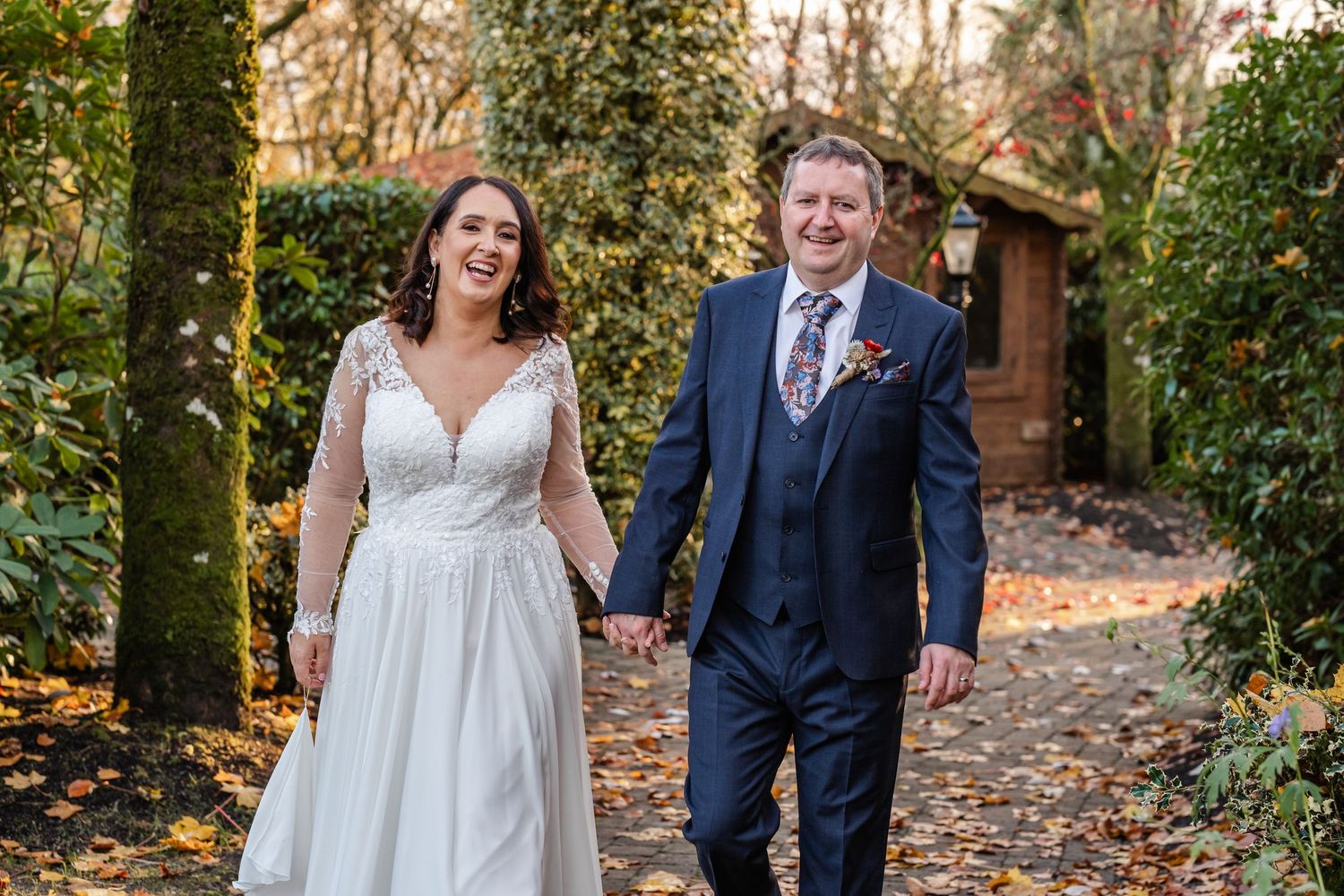 Bride and groom holding hands walking along autumn leaf-covered path outside rustic wedding venue.