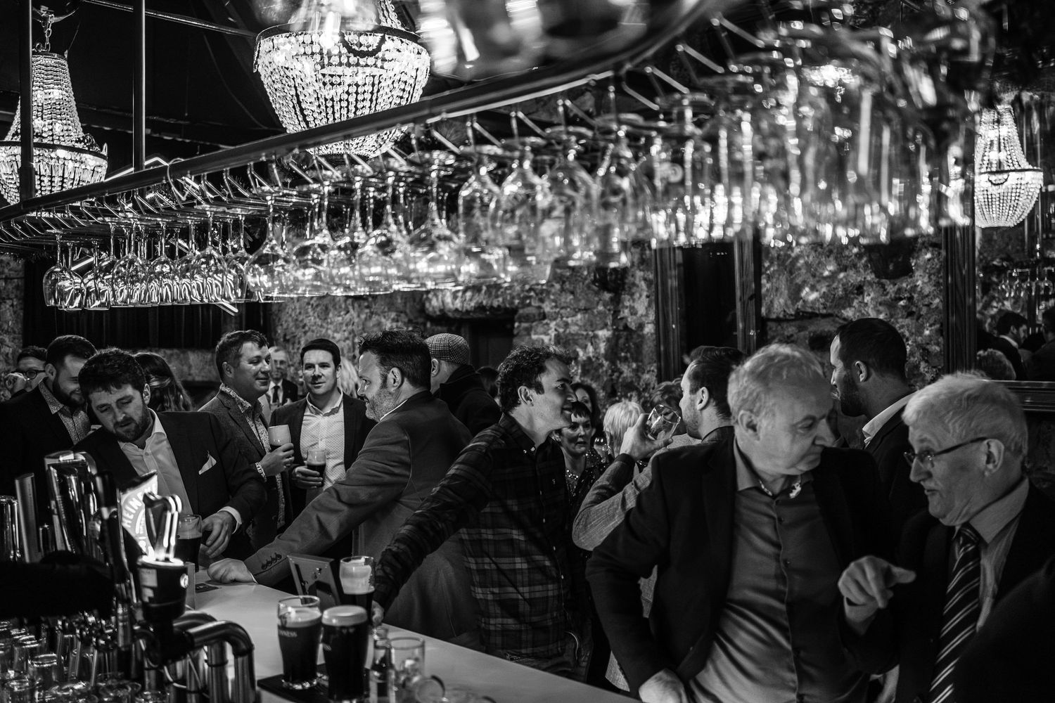Black and white photo of men socializing at a busy upscale bar with crystal chandeliers and hanging wine glasses.