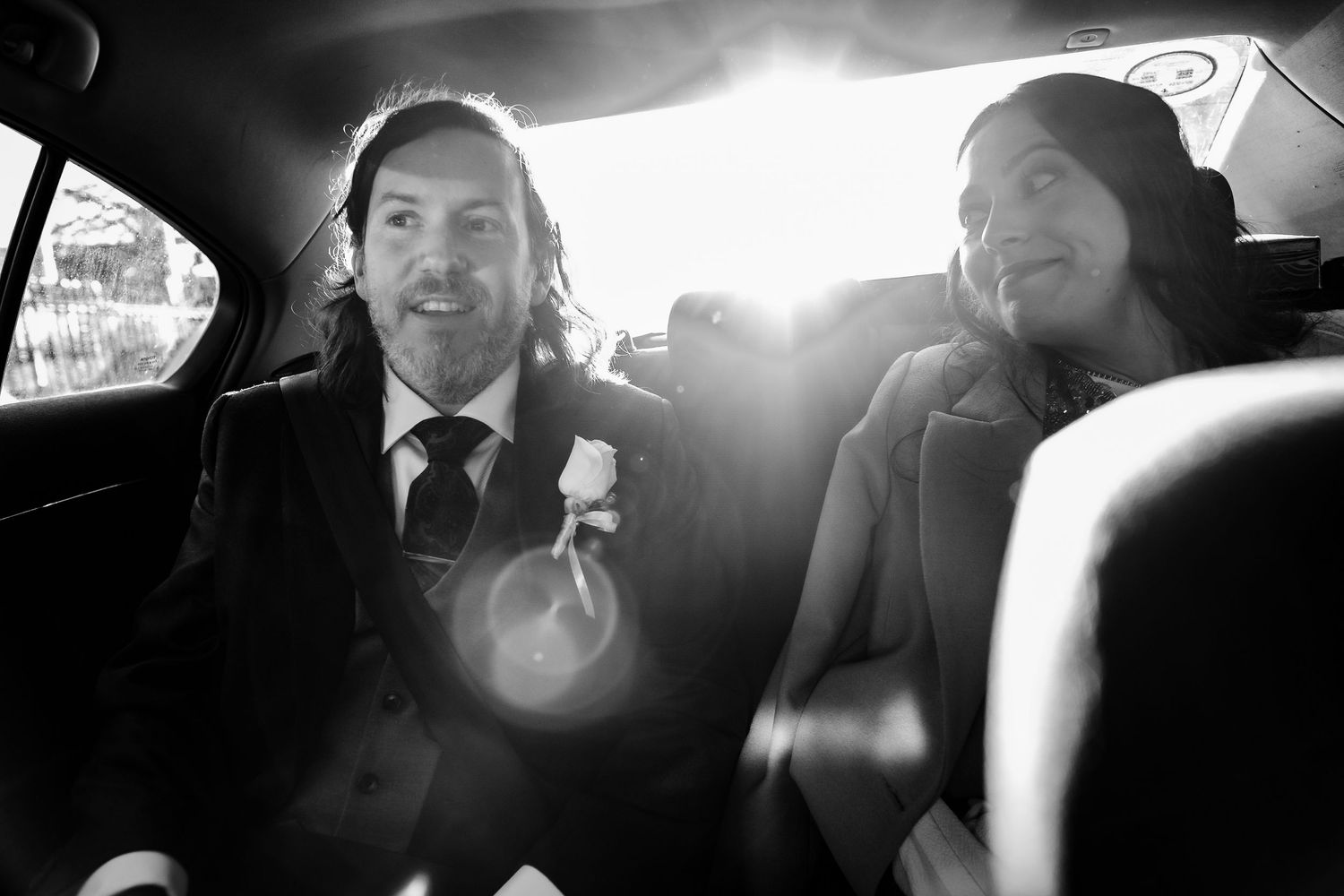 Black and white photo of a bride and groom smiling in the backseat of a car on their wedding day.