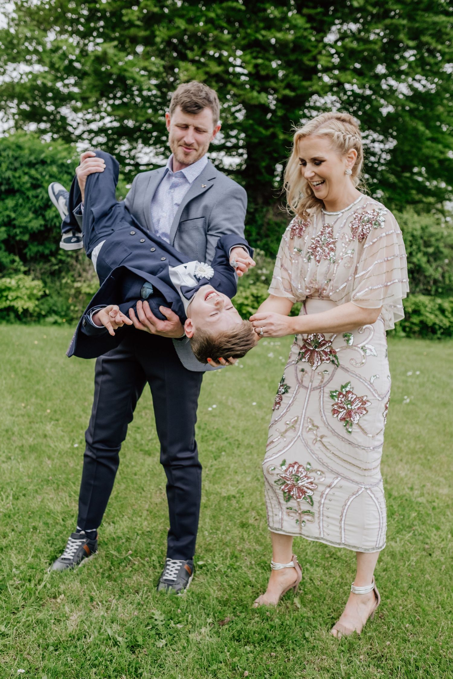 A man and woman in formal attire playfully hold a child upside down on a green lawn at a wedding.