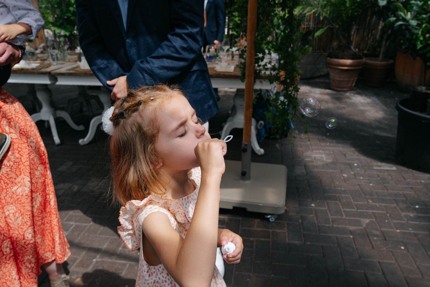 Young girl blowing bubbles outdoors at a garden party on a sunny day.