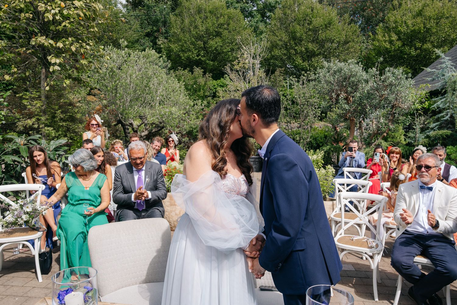Bride and groom share first kiss at outdoor garden wedding ceremony with guests applauding in background.