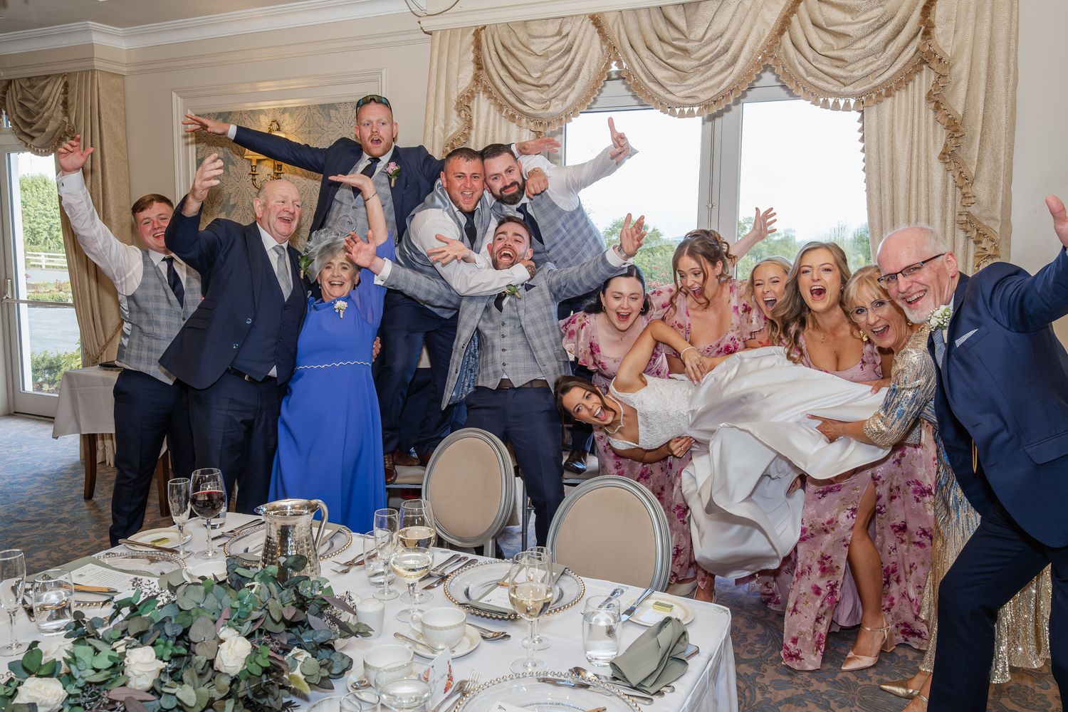 Joyful wedding guests celebrate in elegant reception hall, groom carries bride while group cheers with raised arms.