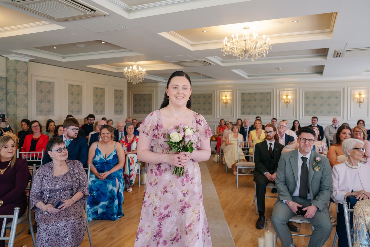 Bridesmaid in floral pink dress walks down aisle holding bouquet at elegant wedding ceremony.