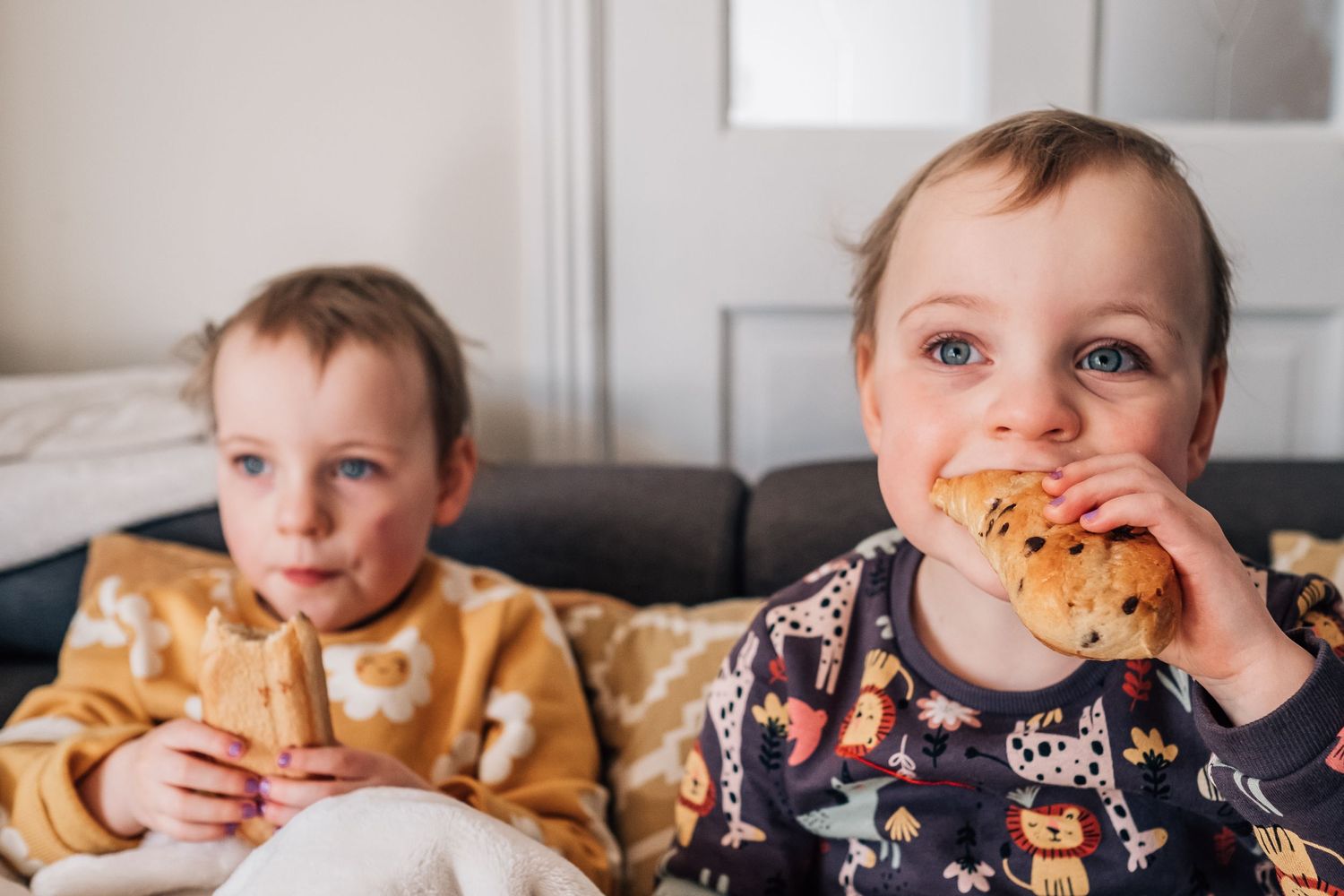 Two children sitting on a couch eating snacks together indoors.