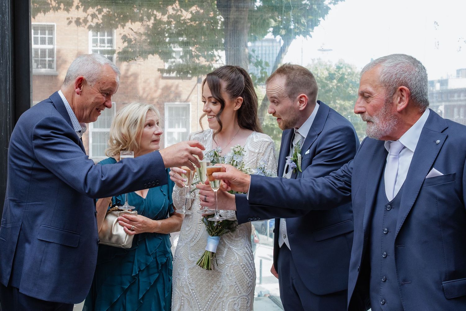 Wedding celebration group toasting with champagne glasses against sunlit windows.