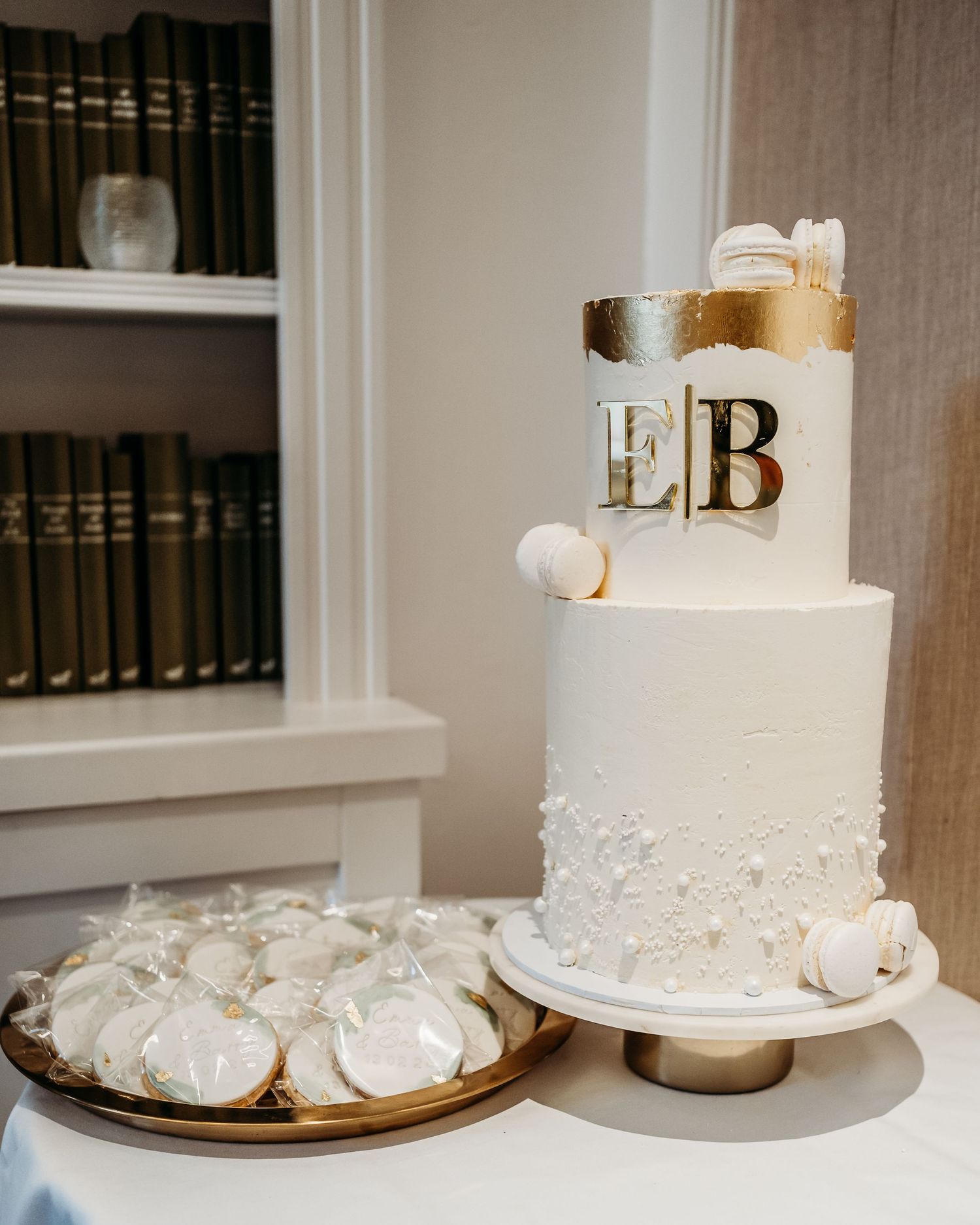 White wedding cake with gold EB monogram and matching white macarons displayed on cake stand.