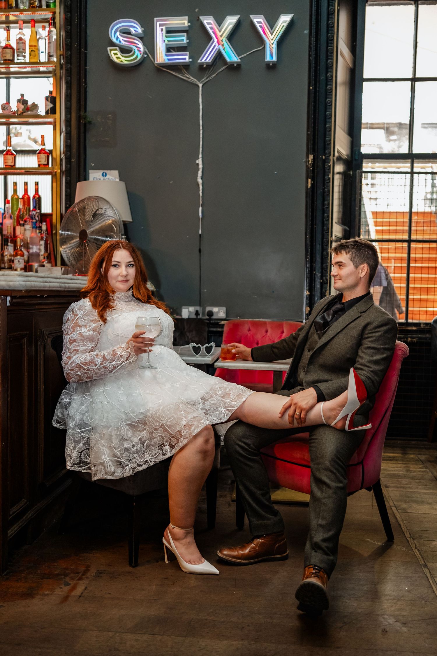 Couple sitting in a bar beneath neon 'SEXY' sign with vintage decor.