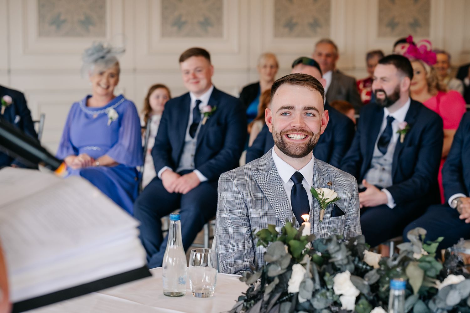 Smiling groom in grey suit seated at wedding ceremony table with guests in formal attire seated behind him.