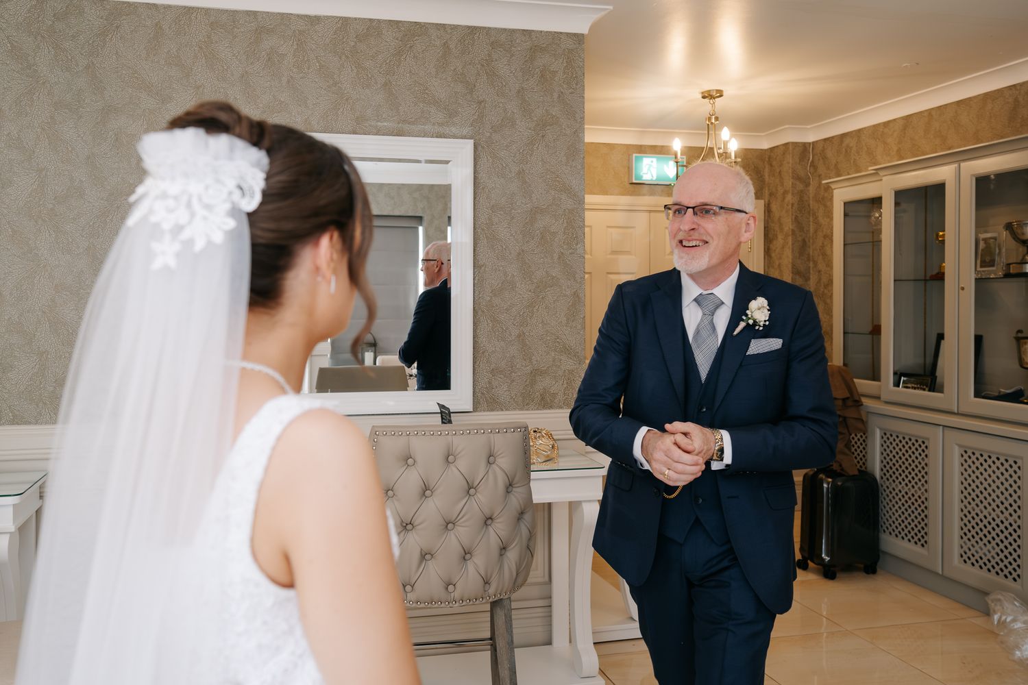 Elderly man in navy suit smiling at bride in white wedding dress and veil during emotional first look moment.