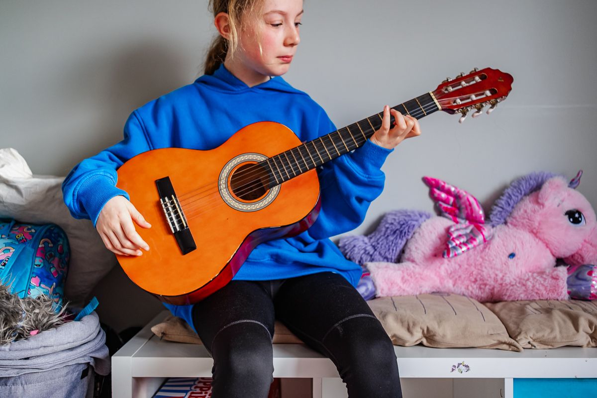 Young girl in blue hoodie playing acoustic guitar seated on bench with stuffed animals.
