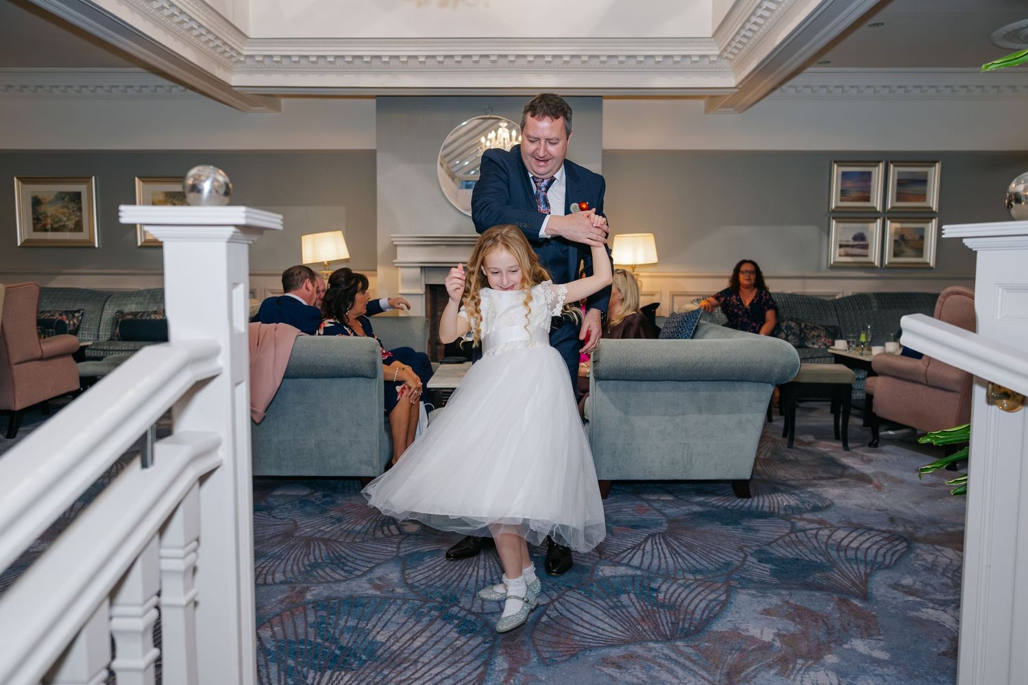 A man in a suit dances joyfully with a young flower girl in a white dress in an elegant hotel lobby.