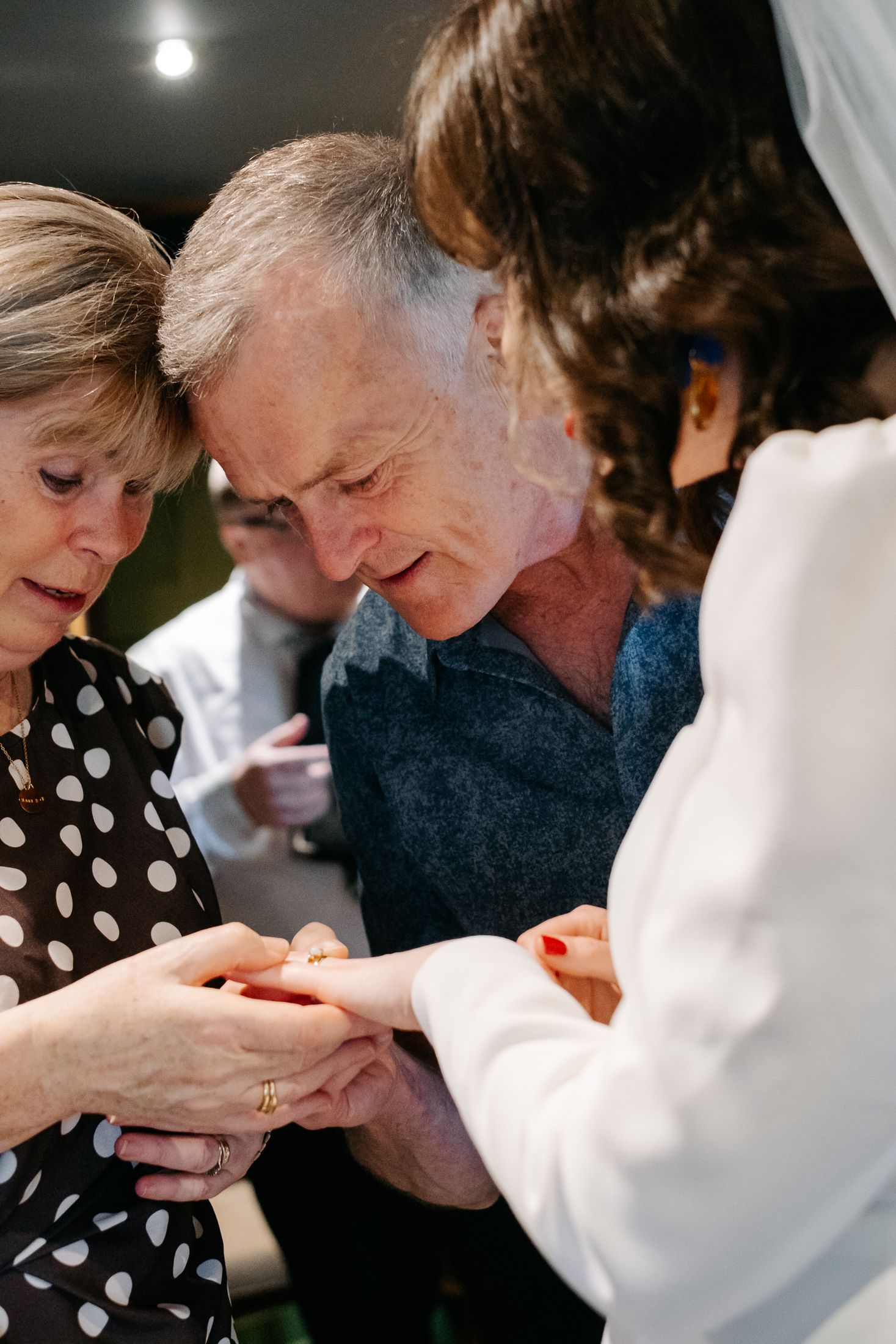 Elderly couple examining a wedding ring during a ceremony, with a bride in white veil extending her hand.