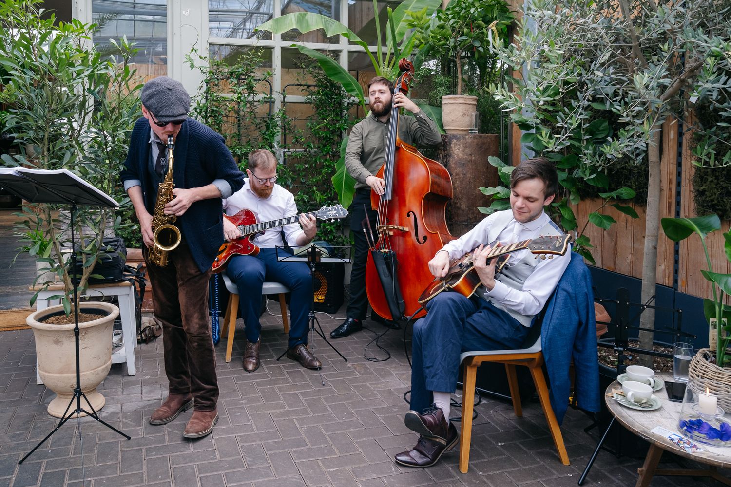 Jazz quartet performing outdoors with saxophone, guitars, and double bass amid lush green plants.