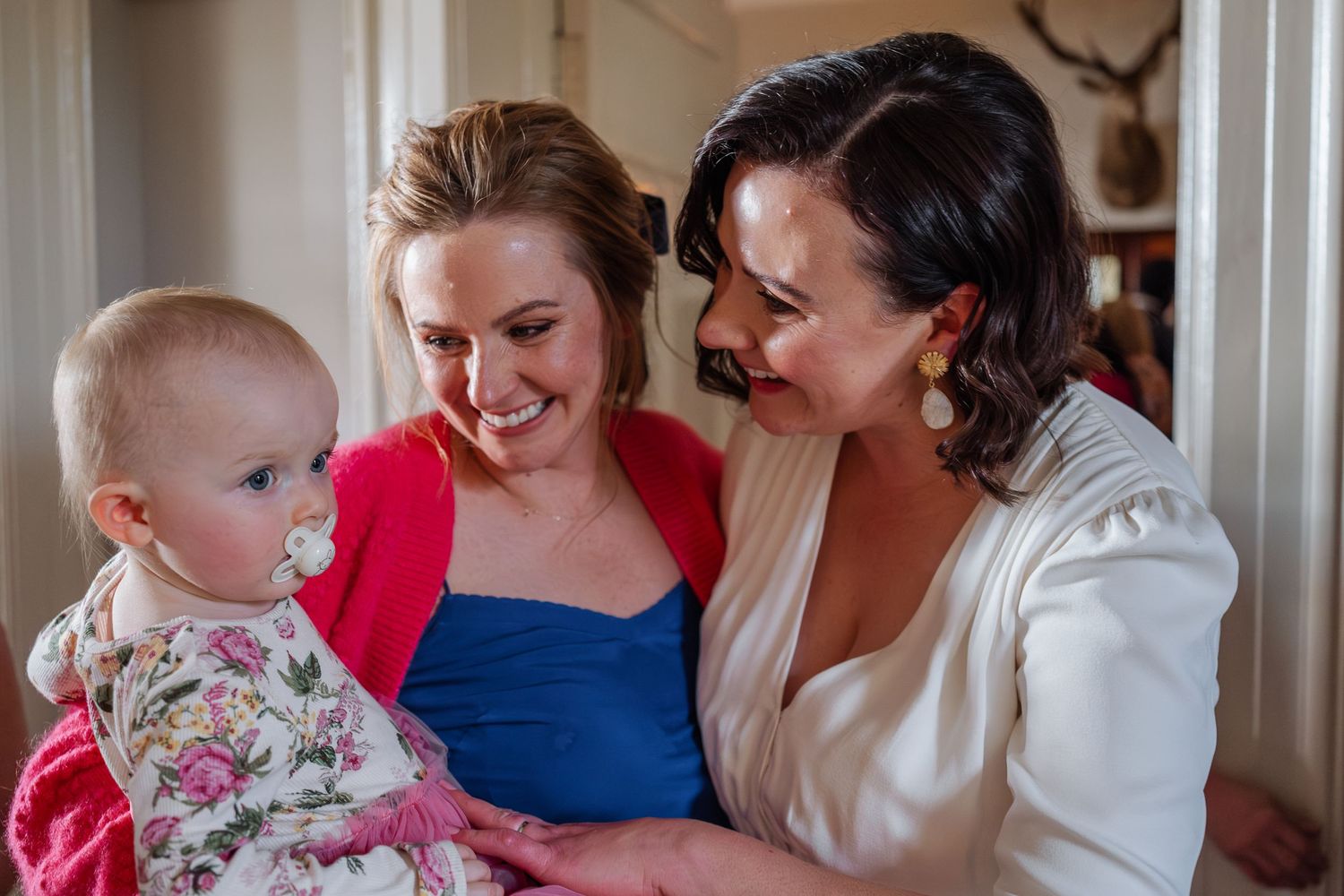 Two women smiling at a baby in floral outfit, sharing a joyful moment at a wedding celebration.