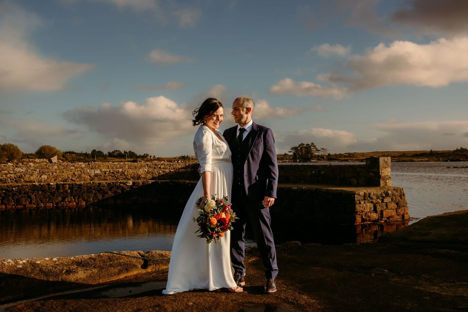 Bride and groom share a romantic moment by a waterfront, she holds colorful bouquet, he wears navy suit, golden sunset light.