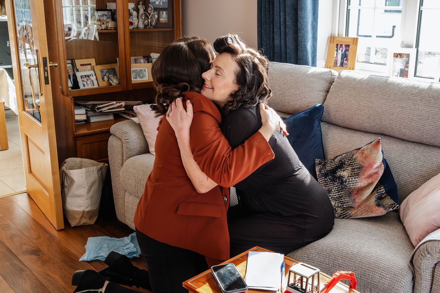 Two women share a warm embrace on a sofa during a wedding preparation at home.