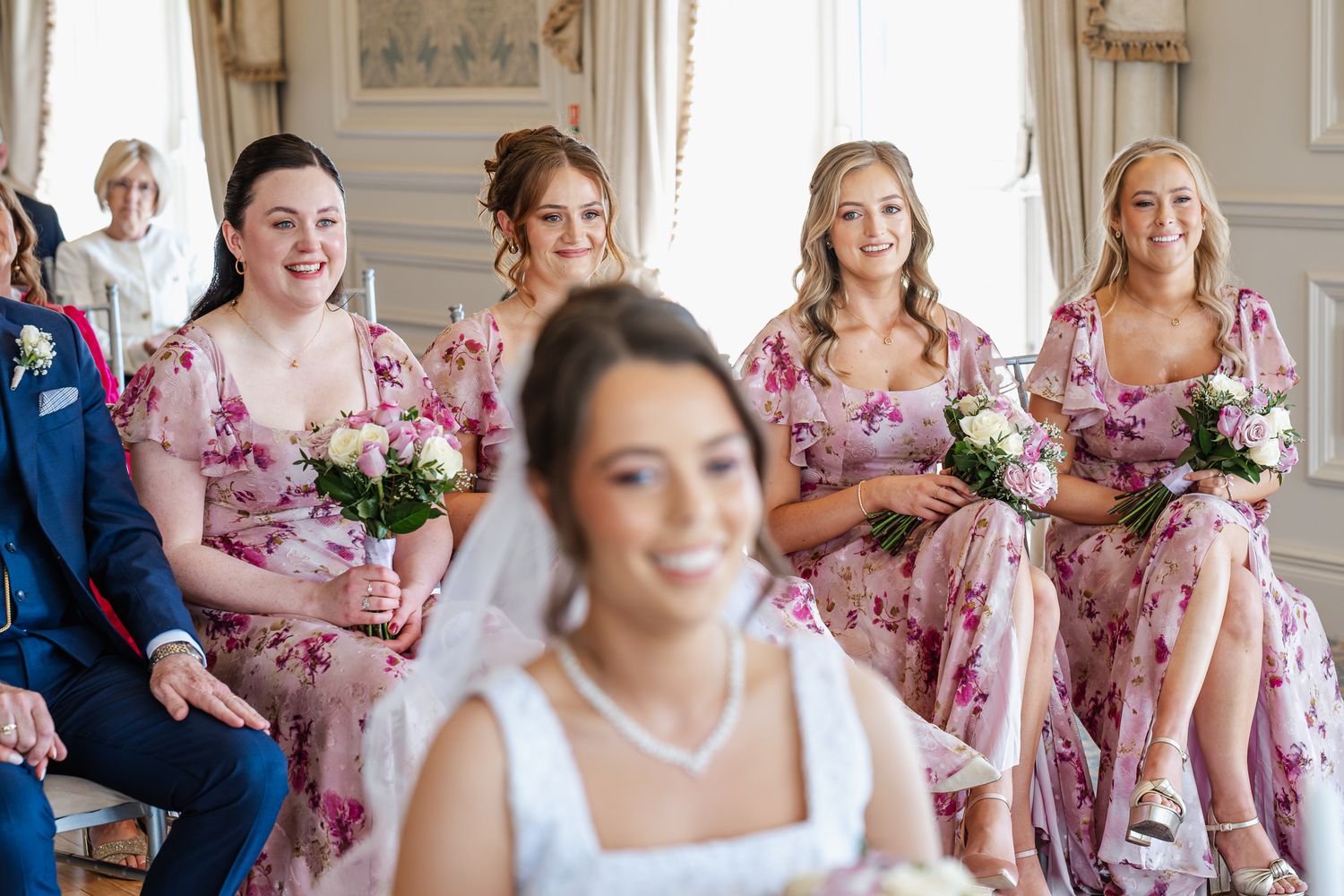 Bride in white veil smiles as bridesmaids in pink floral dresses hold bouquets at an elegant wedding ceremony.