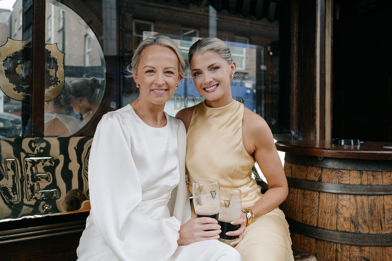 Two women in elegant wedding attire smiling together outside a pub, holding Guinness glasses.