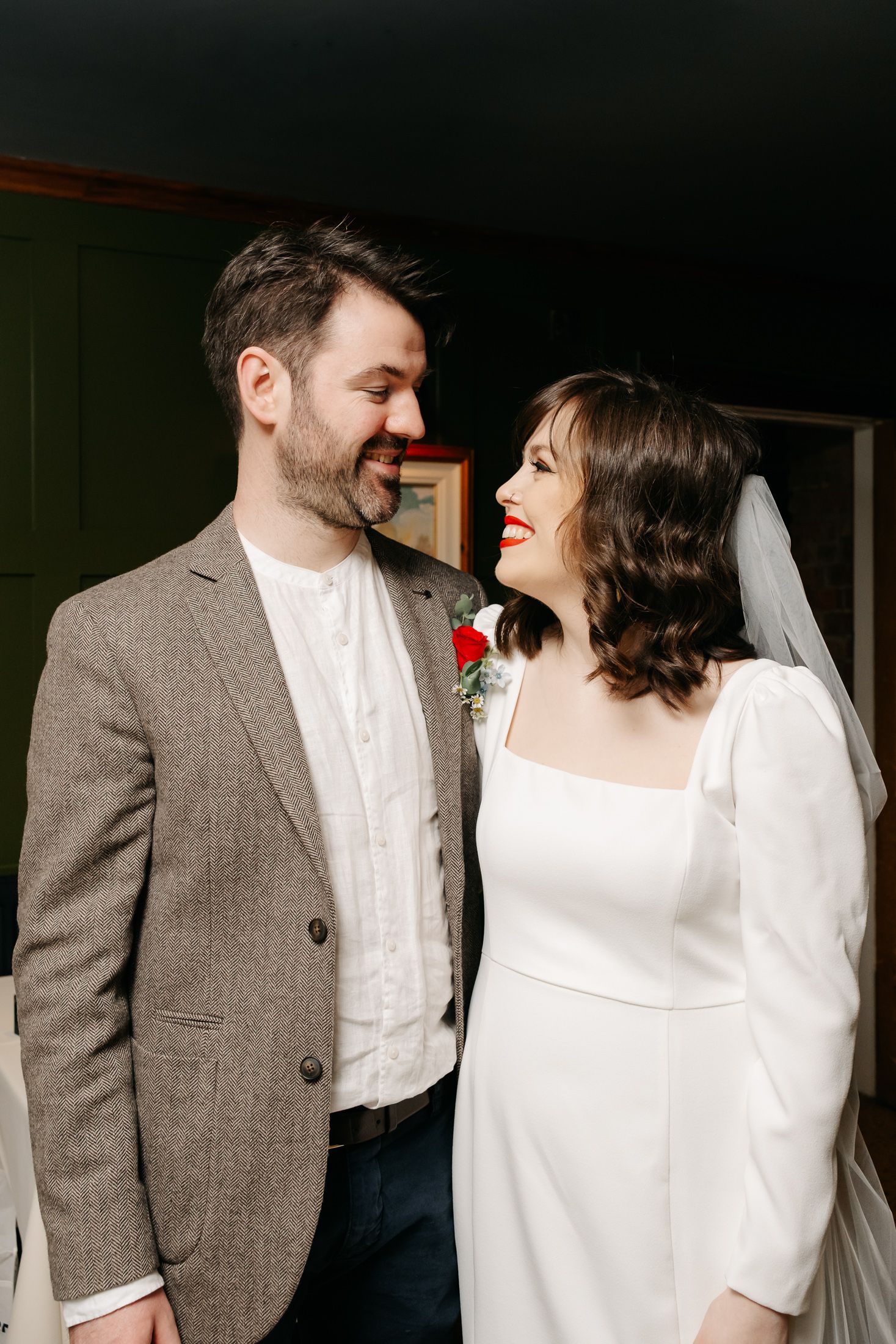 A smiling bride in a white dress and veil with red lips gazes at her bearded groom in a gray tweed suit.