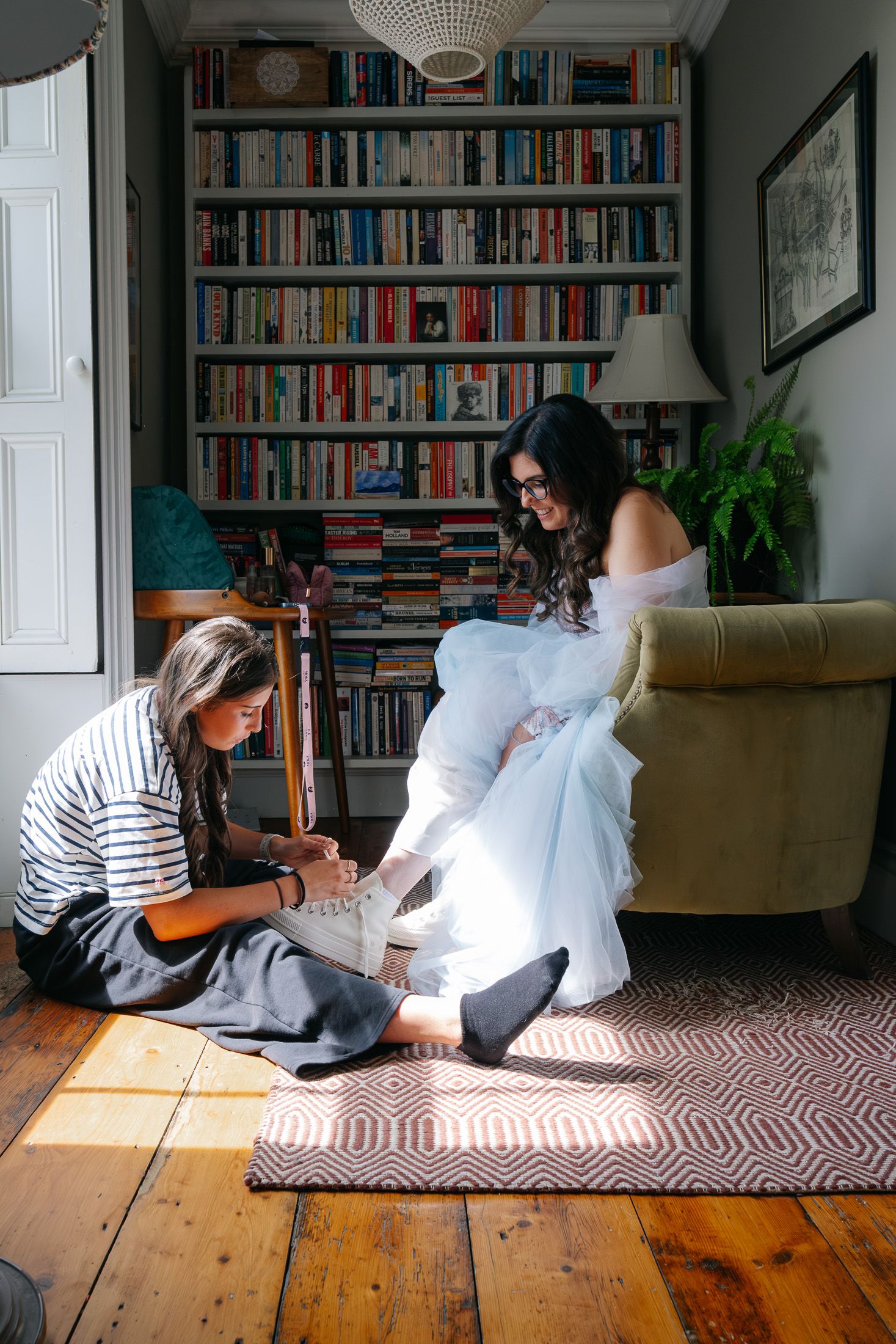Bride in white dress getting shoes tied by bridesmaid in striped top, seated by bookshelf in cozy room.