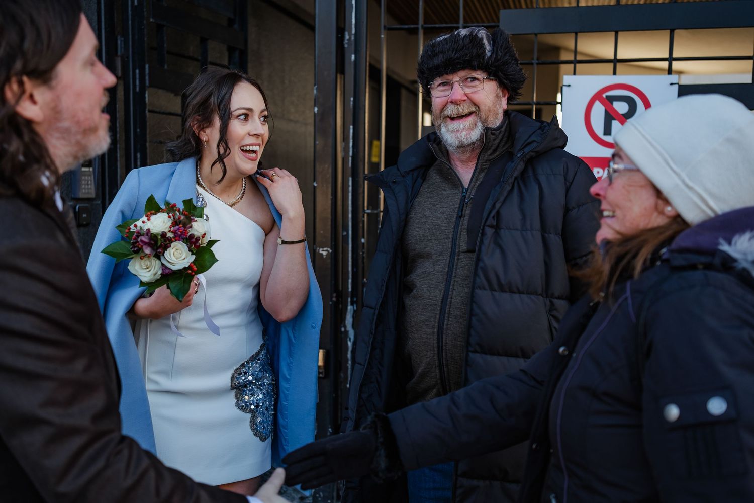 Bride in white dress holding bouquet chats with guests outside in winter, wearing blue wrap, near no parking sign.
